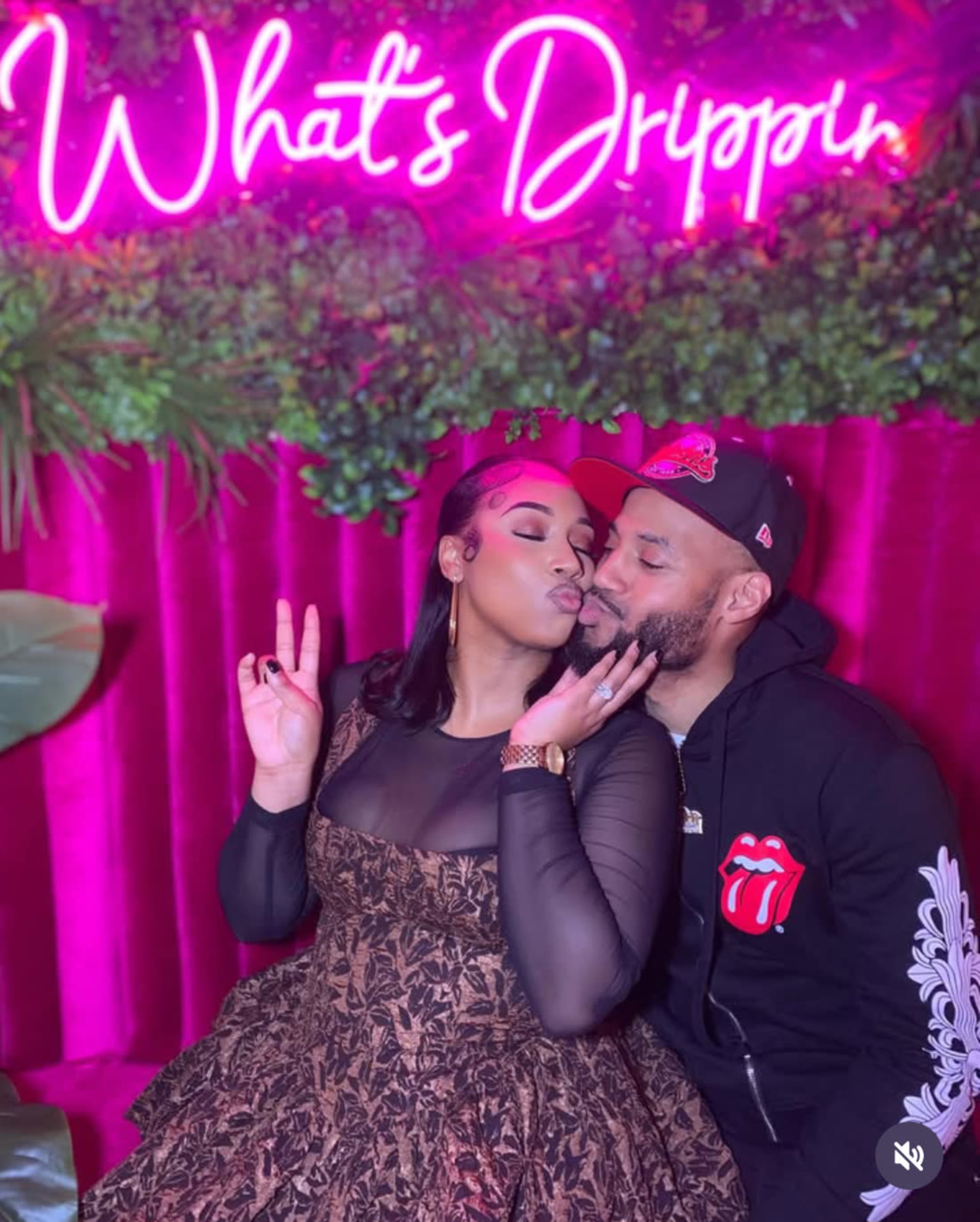 A couple poses playfully in front of a neon sign that reads "What's Drippin" against a backdrop of plants and pink fabric.