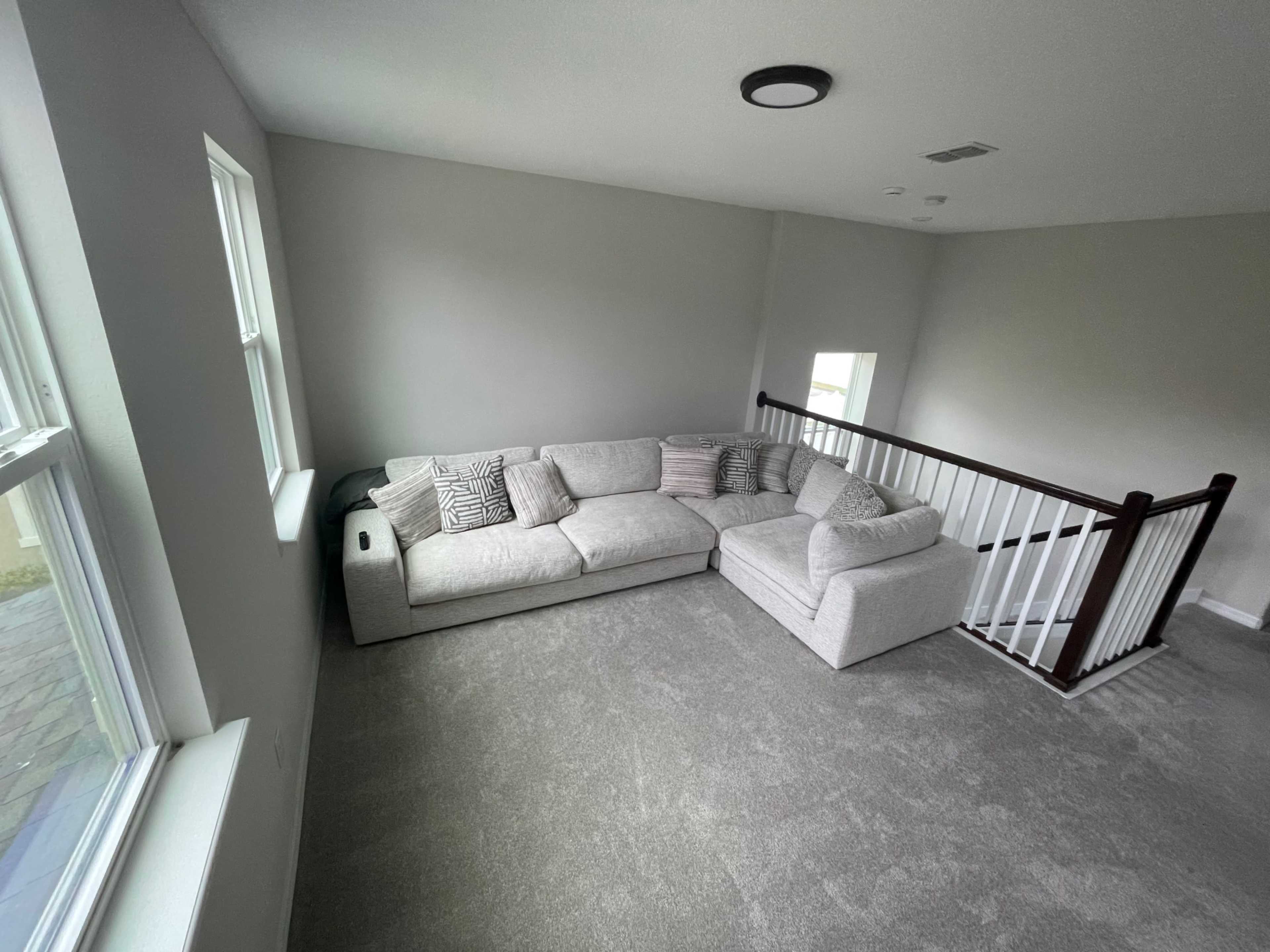 A light gray sectional sofa with decorative pillows is positioned in a well-lit room featuring two windows and a staircase railing.
