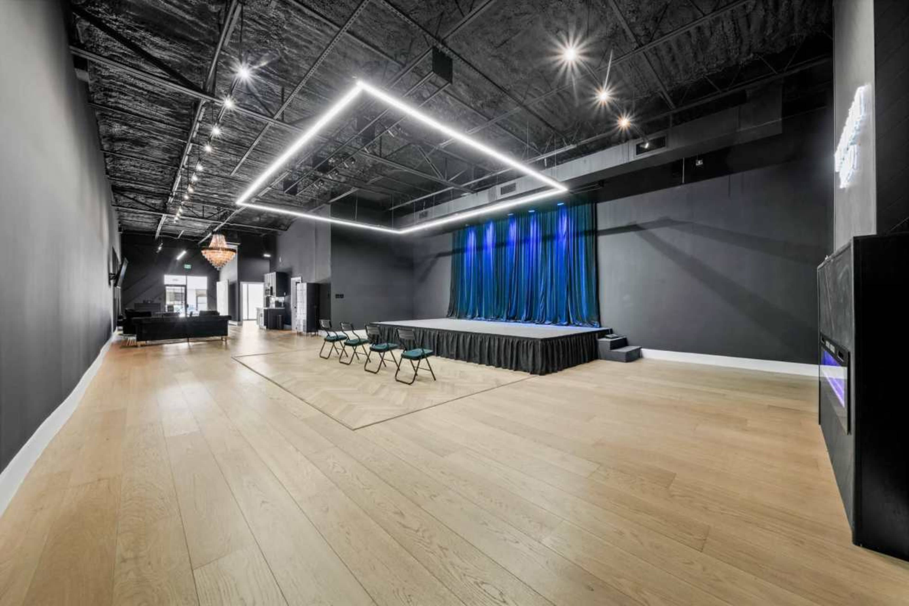 The image shows an empty performance space with a stage at one end, lined with blue curtains, and a row of chairs set up in front.