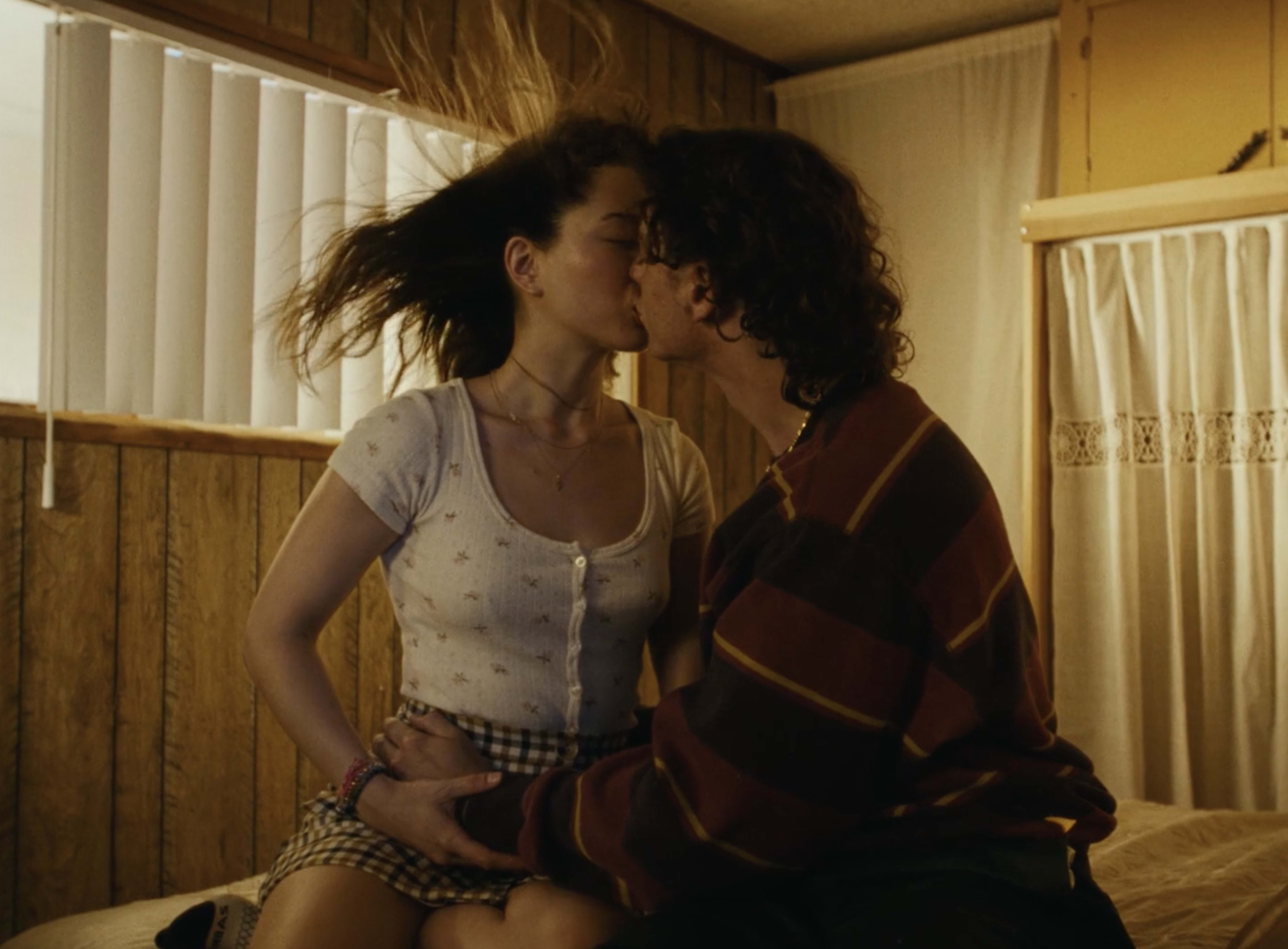 A couple is kissing on a bed in a room with wooden walls and natural light coming through the blinds.