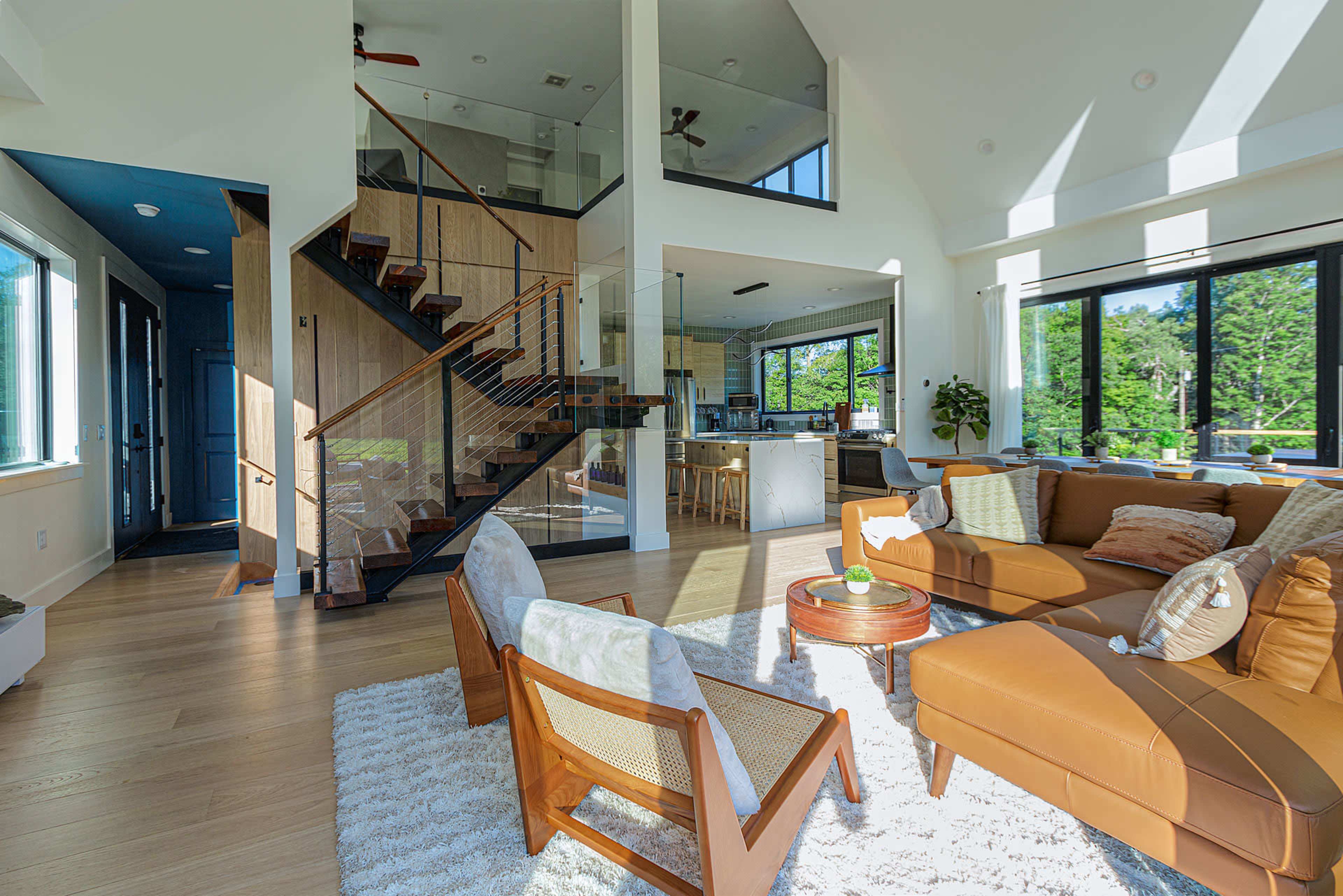 A modern living room with a brown leather sofa, two armchairs, and a staircase leading to an upper level, all illuminated by natural light from large windows.