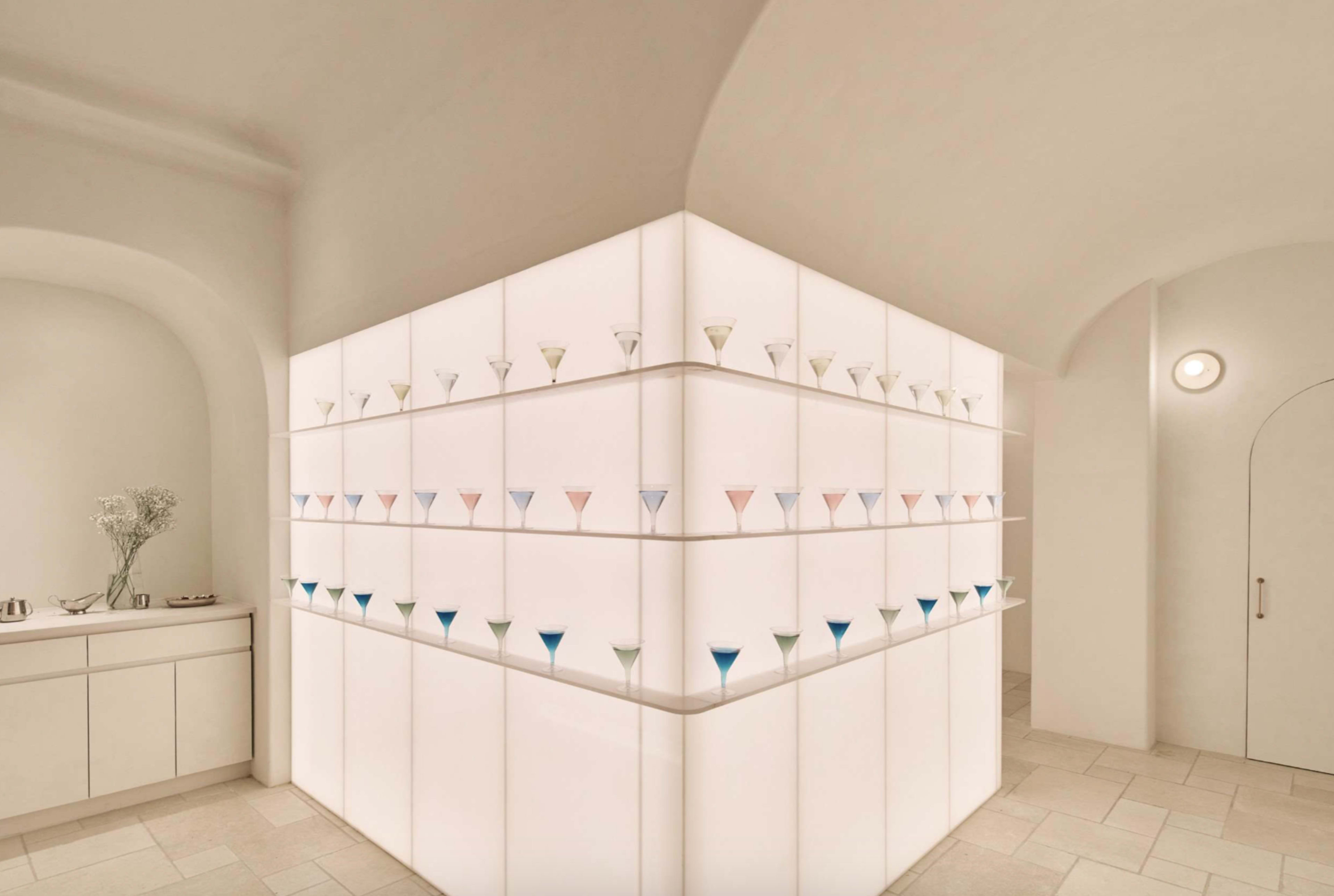 The image depicts a modern interior space featuring a corner wall with multiple shelves holding colorful glass vessels arranged systematically.