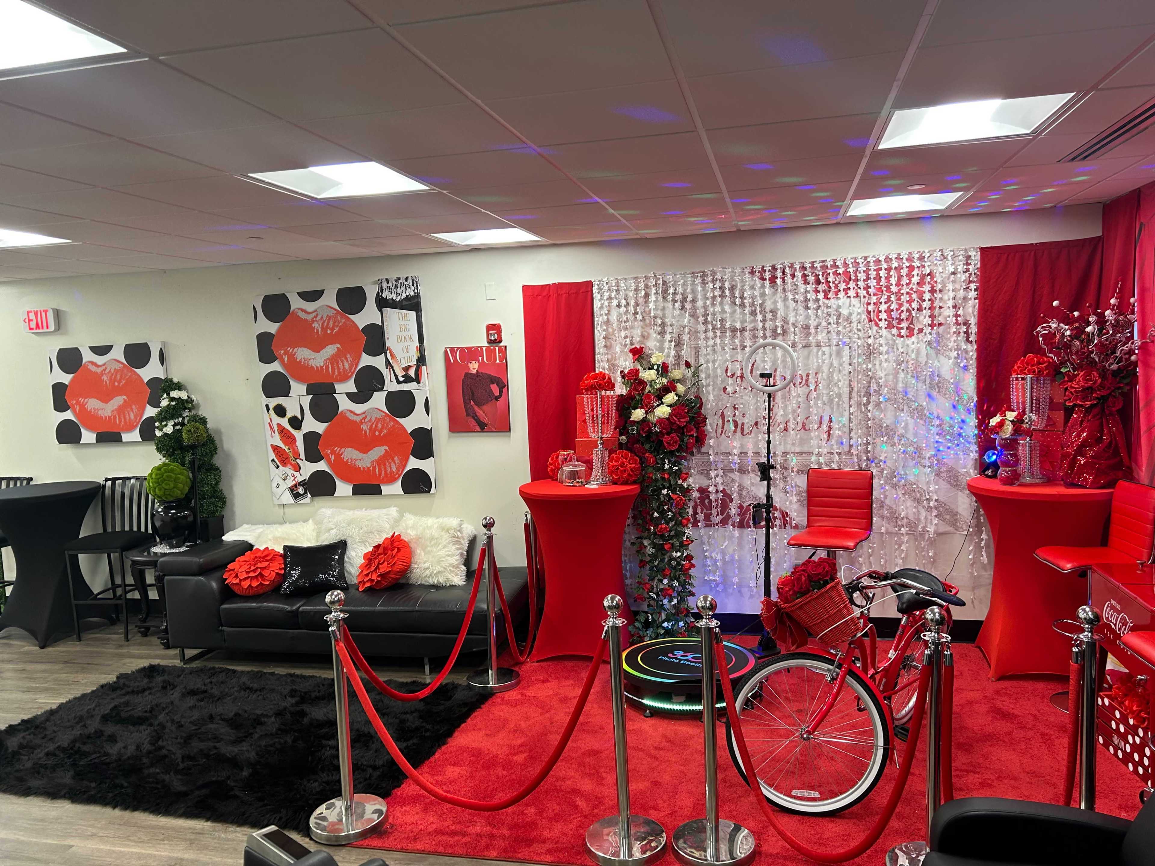 A decorated interior space featuring red and black decor, including a couch, two high tables, floral arrangements, and a bicycle with a vibrant backdrop.
