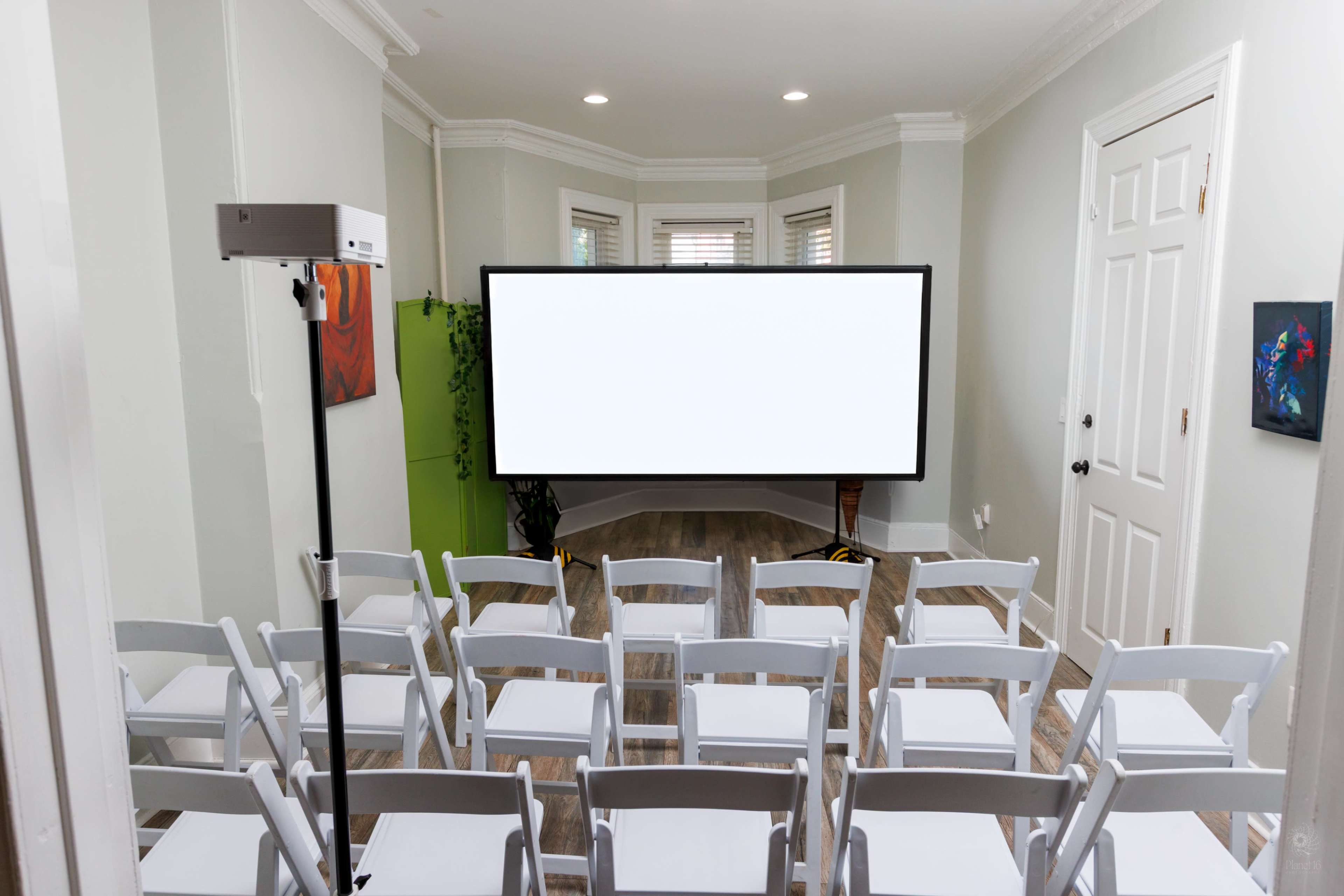 The image shows a room set up with white folding chairs facing a large blank screen, with a projector on a stand nearby.