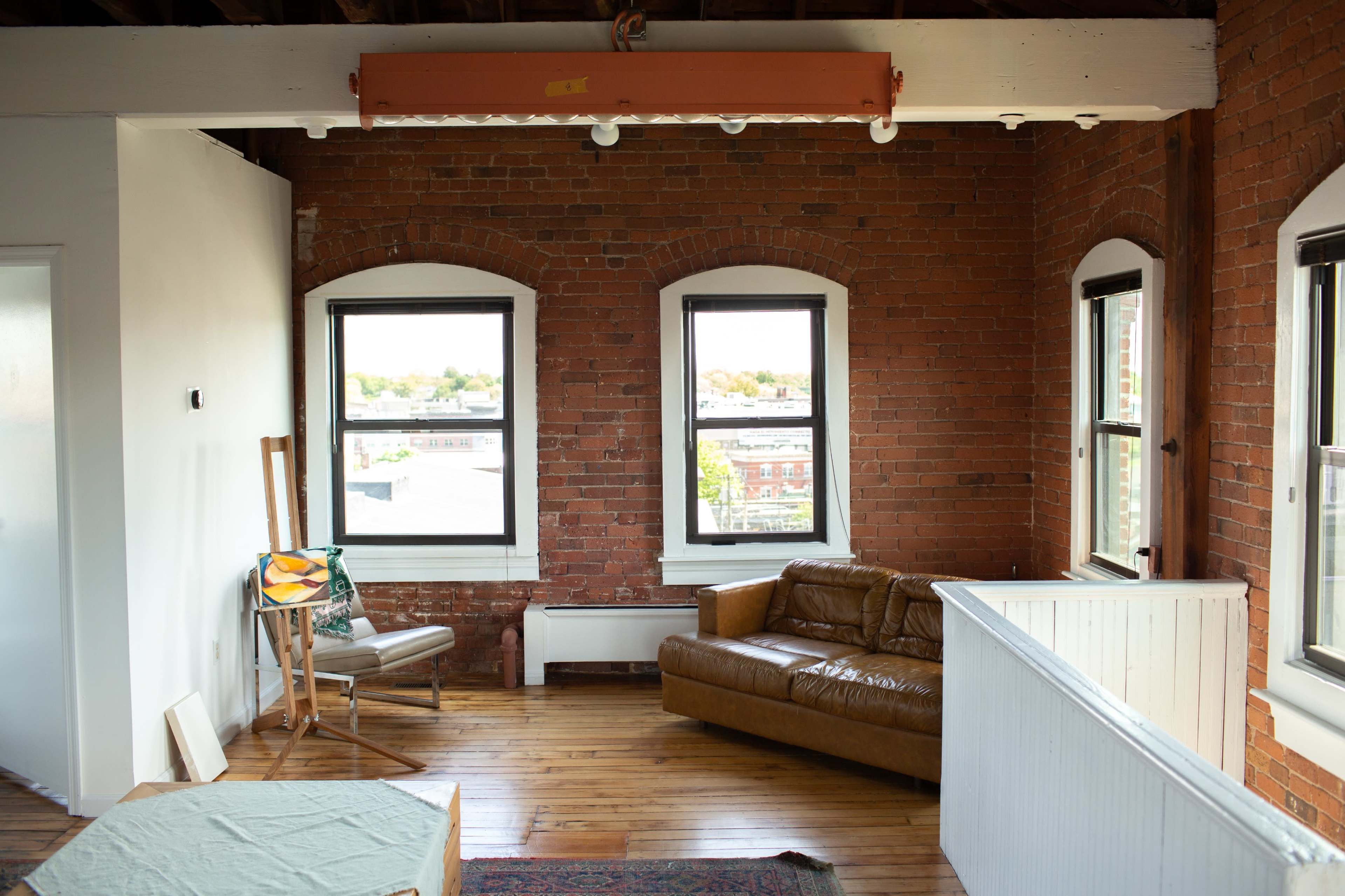 A bright, spacious room with exposed brick walls, large windows, a brown leather couch, and wooden flooring.