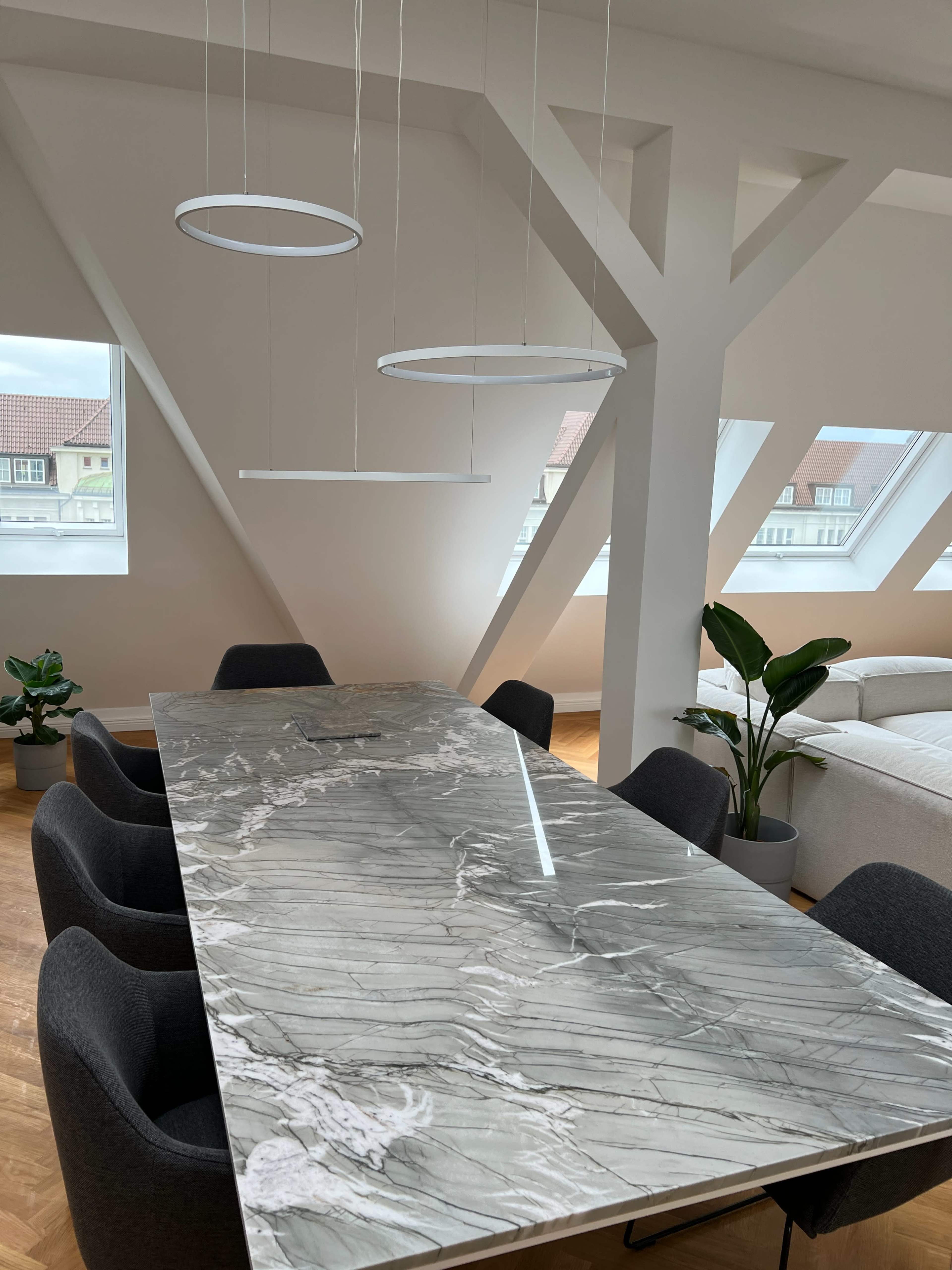 A long marble dining table is surrounded by black chairs in a well-lit room with modern lighting fixtures and large windows.