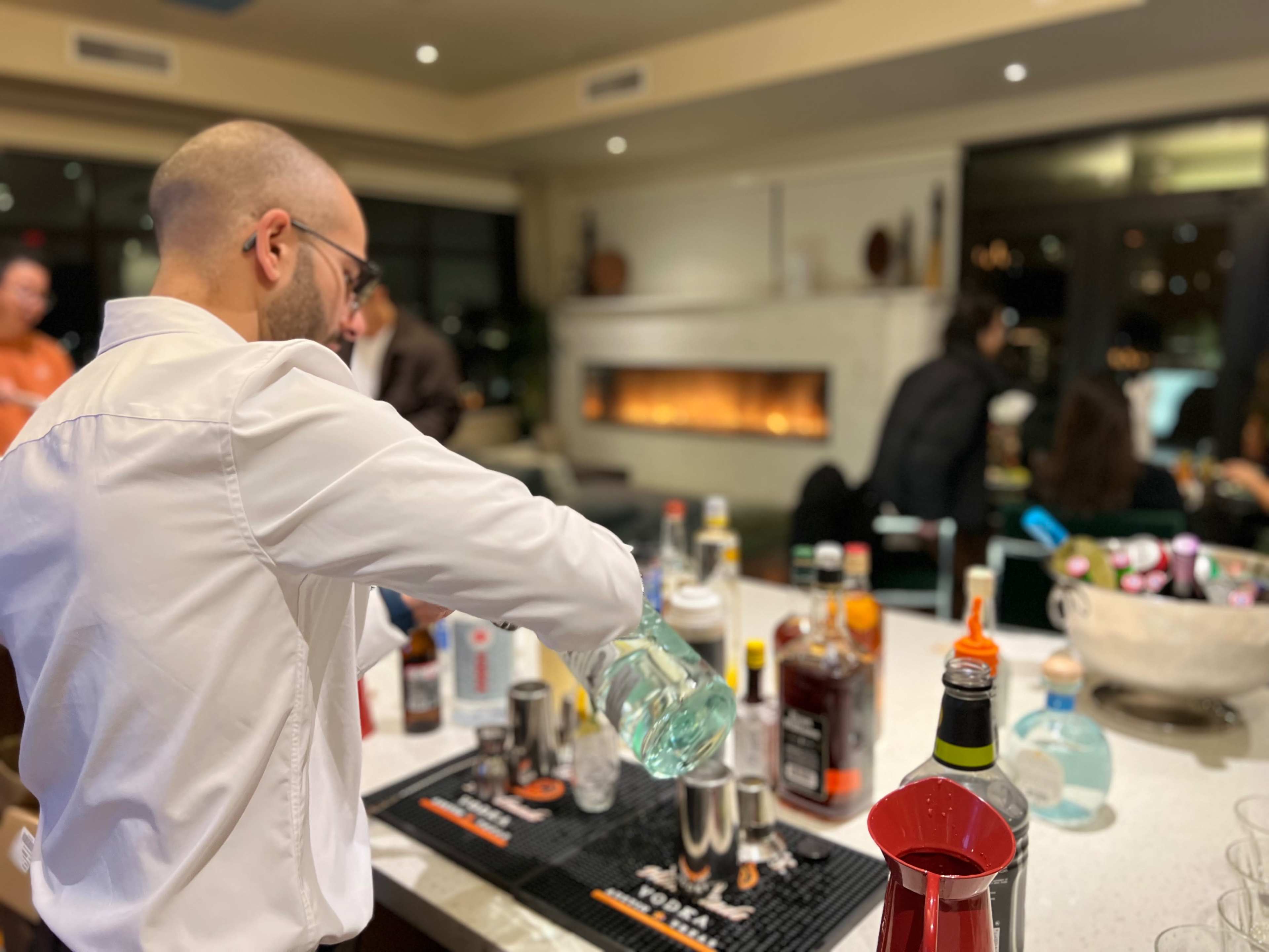 A bartender in a white shirt pours a clear liquid from a bottle into a cocktail shaker at a bar filled with various liquor bottles.