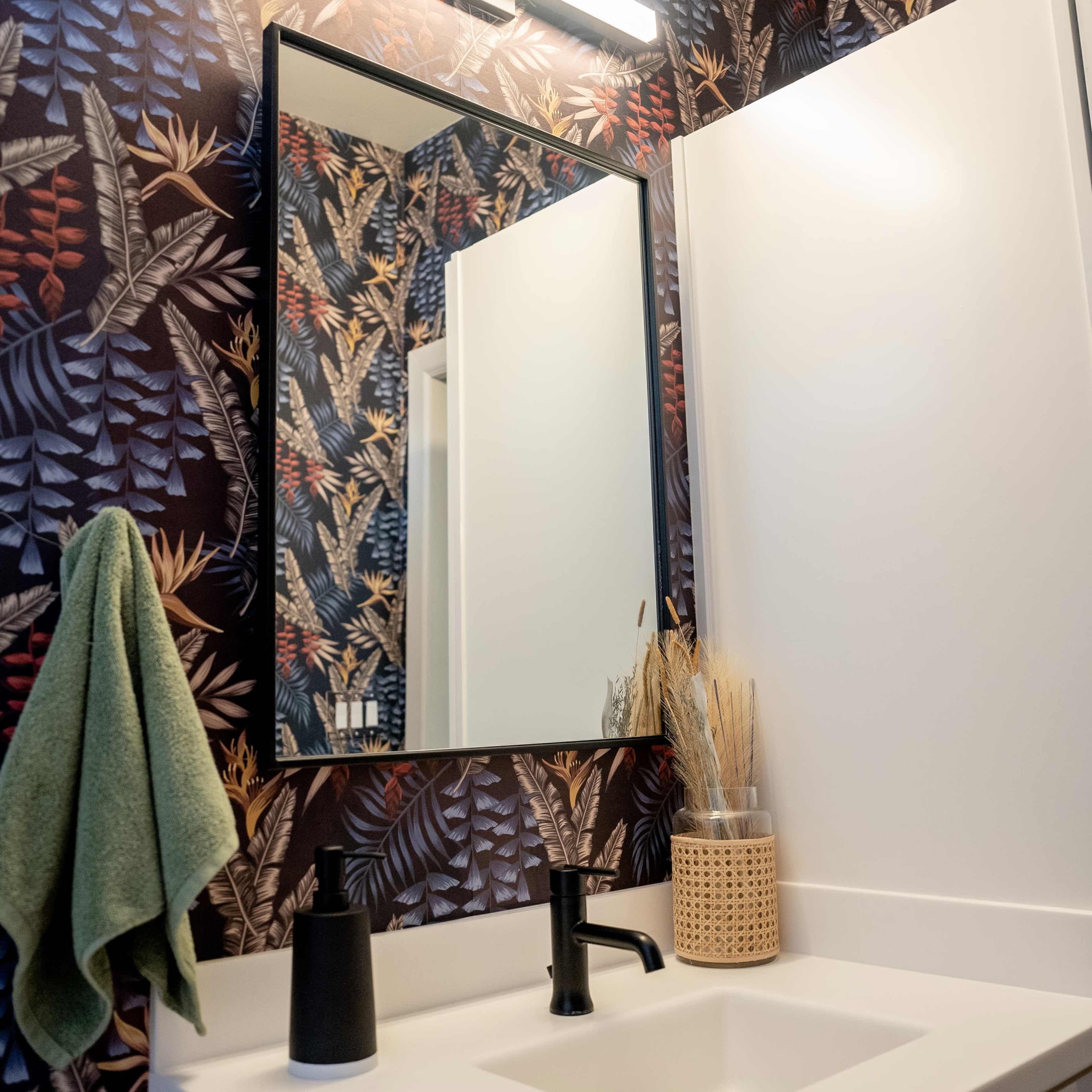 A bathroom scene features a large mirror above a white sink, surrounded by floral-patterned wallpaper and accompanied by a green towel and decorative items.