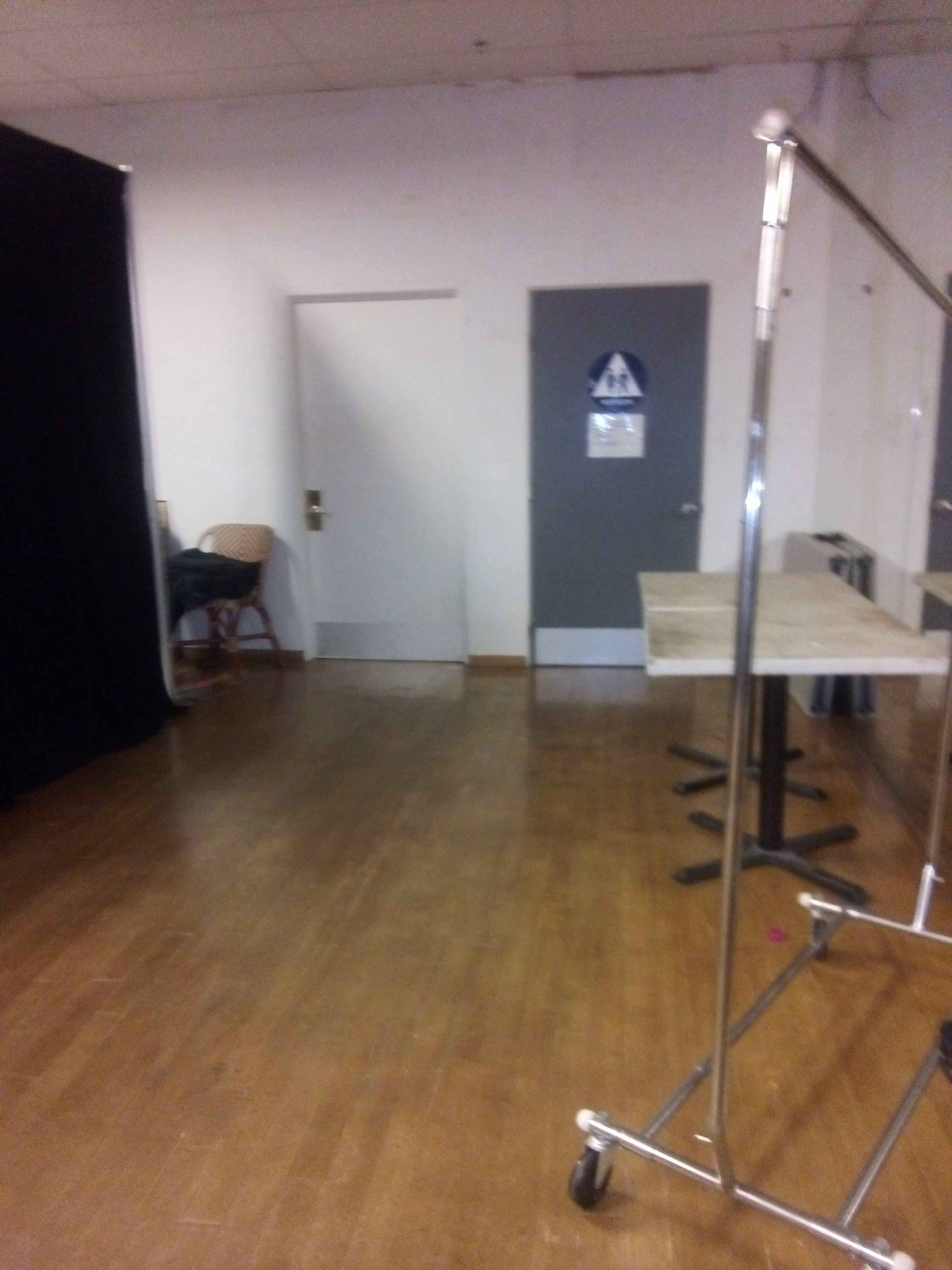 A room with a wooden floor, a closed door labeled as a restroom, a folding chair, and a clothing rack near a black curtain.