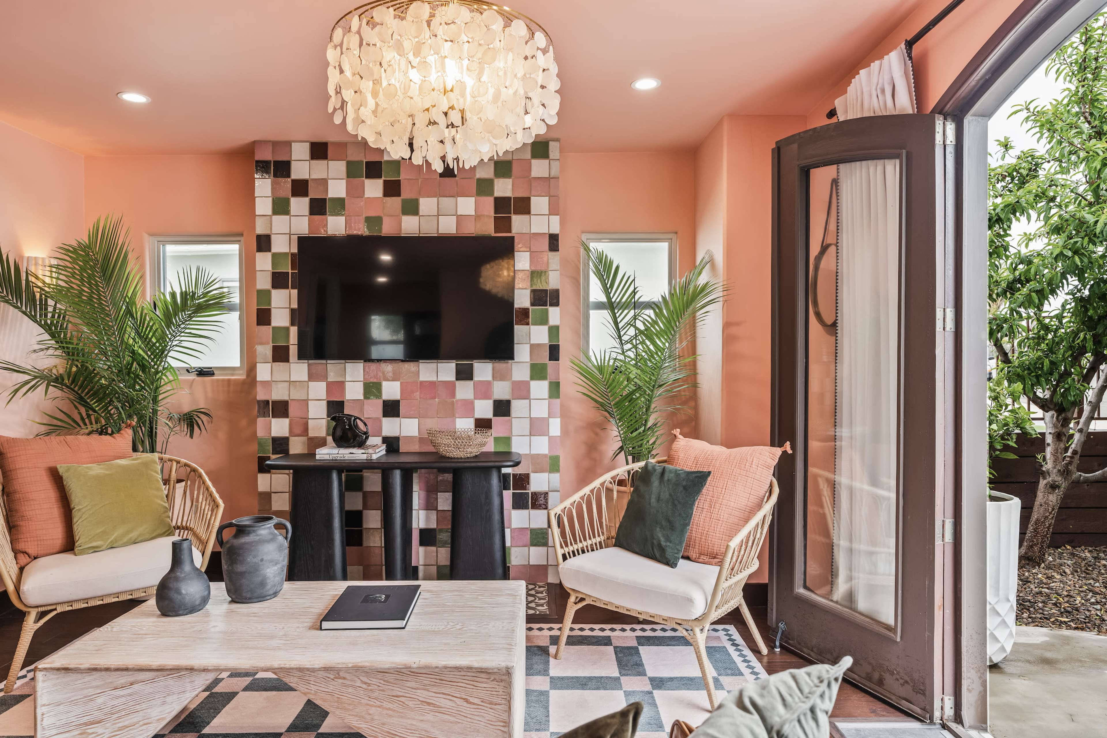 A cozy living space features a light pink wall, a patterned tile fireplace, a mounted TV, and two wicker chairs with decorative cushions surrounding a wooden coffee table.