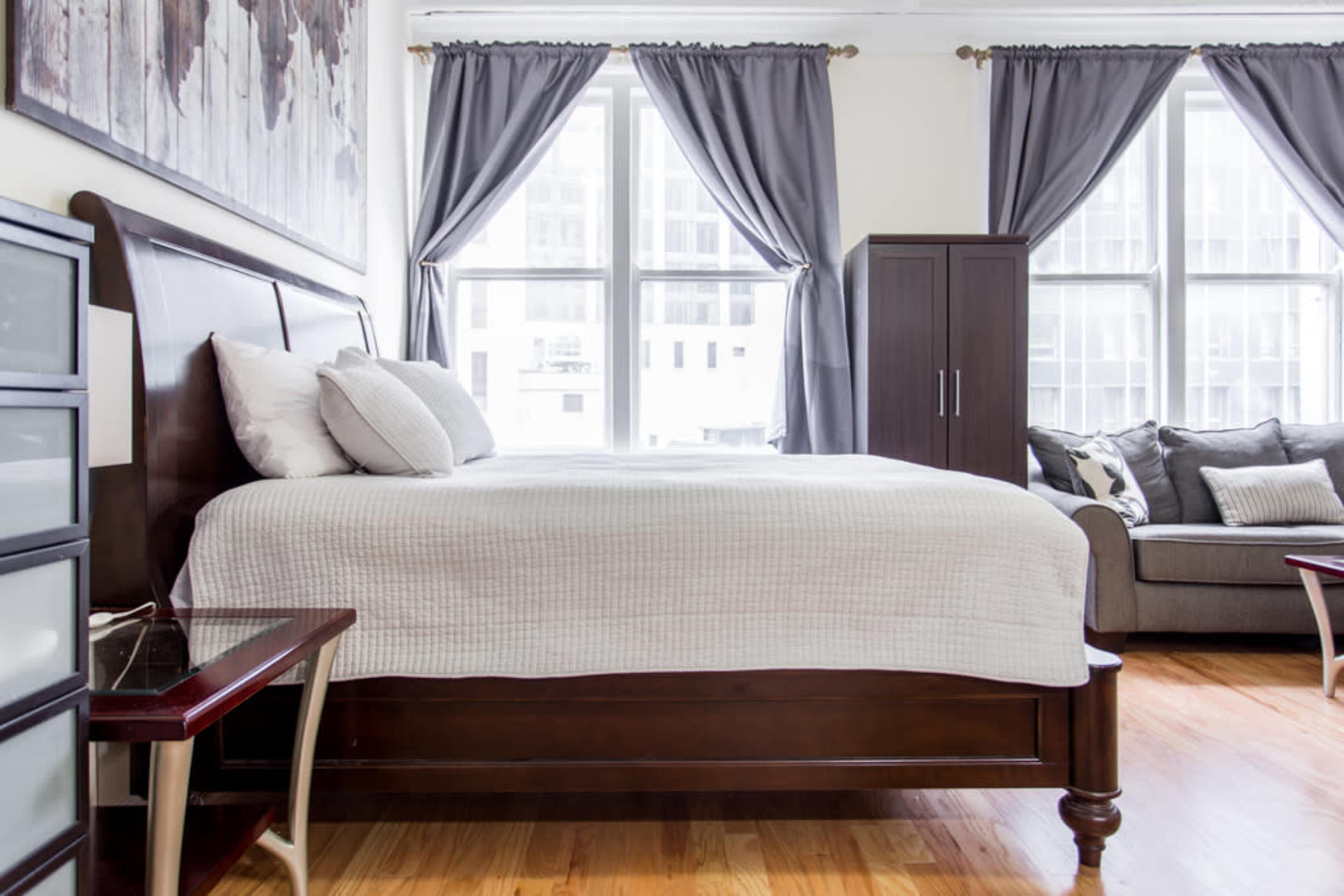 A bedroom with a wooden bed, gray curtains, a small side table, and a gray sofa.