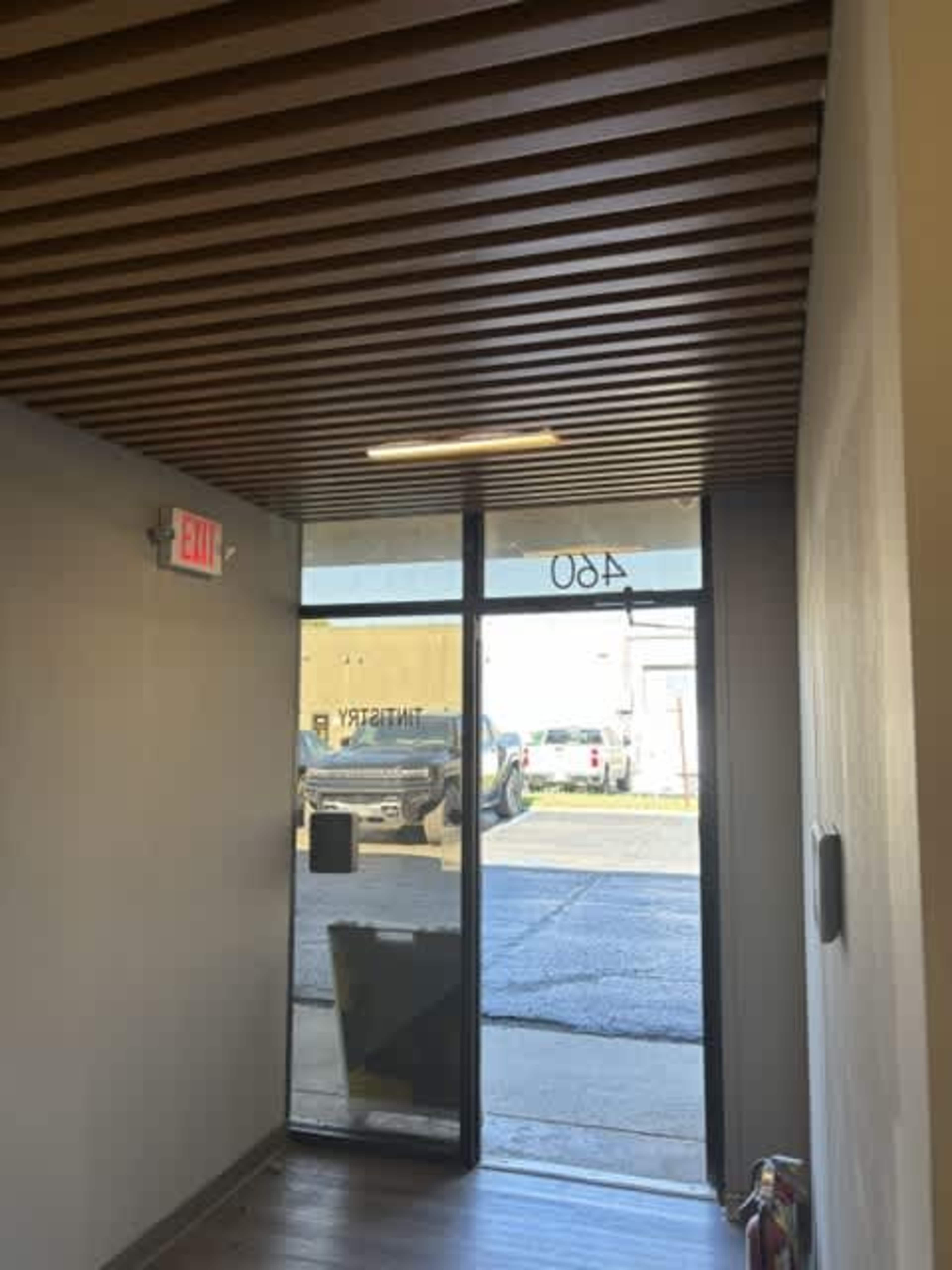 The image depicts a hallway with wooden ceiling slats, leading to a glass door that opens to an exterior view of a parking area.