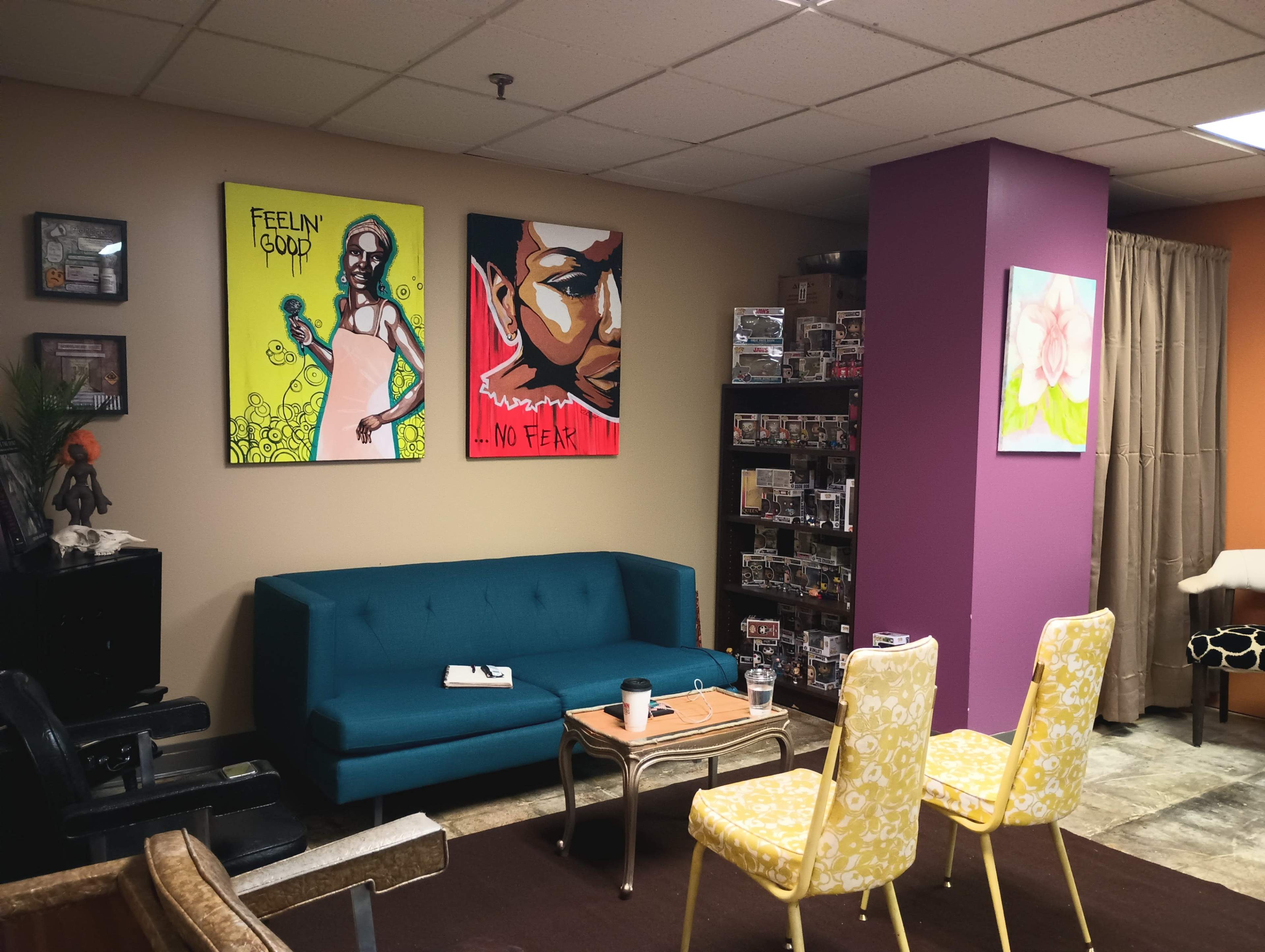 The image shows a small, colorful waiting room featuring a blue couch, two yellow chairs, and various artworks on the walls.