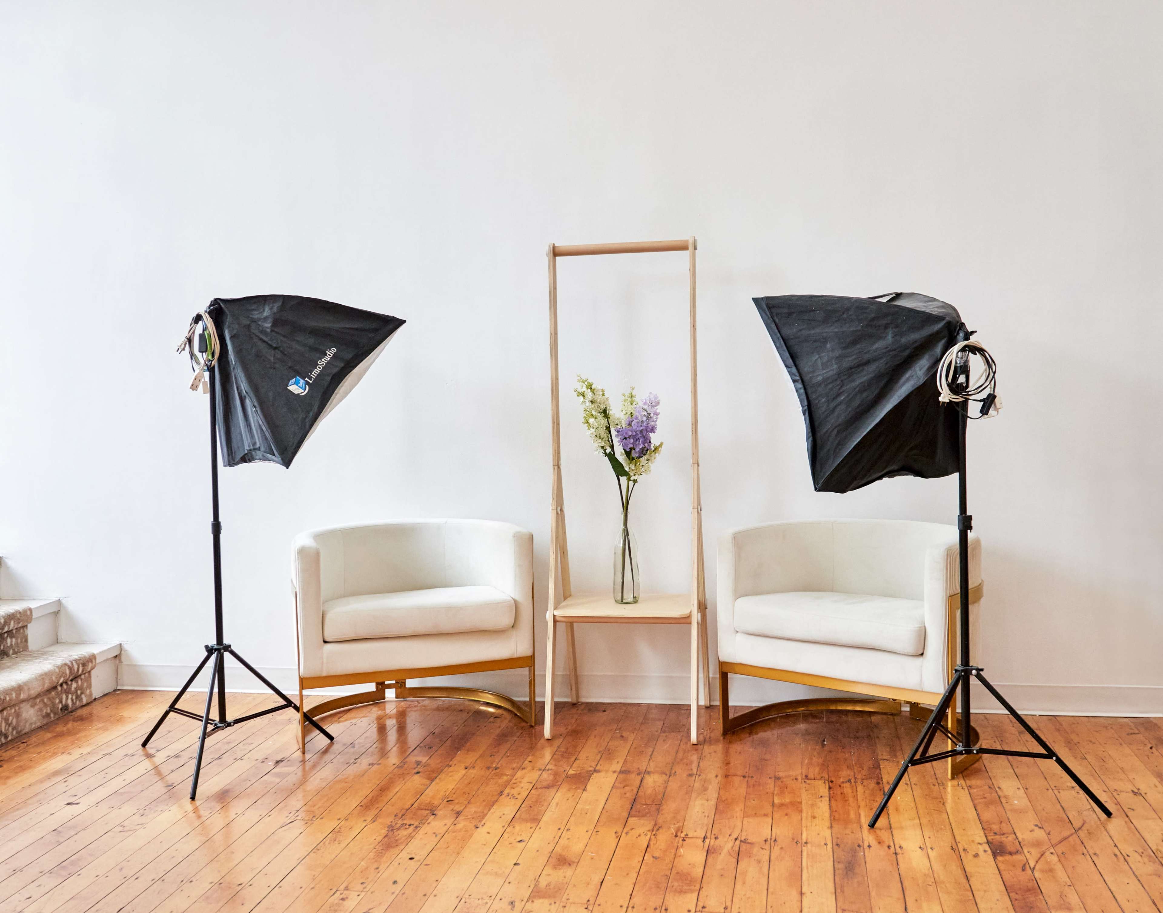 Two white chairs positioned on a wooden floor, with a simple wooden stand holding a vase of flowers between two studio lighting setups.