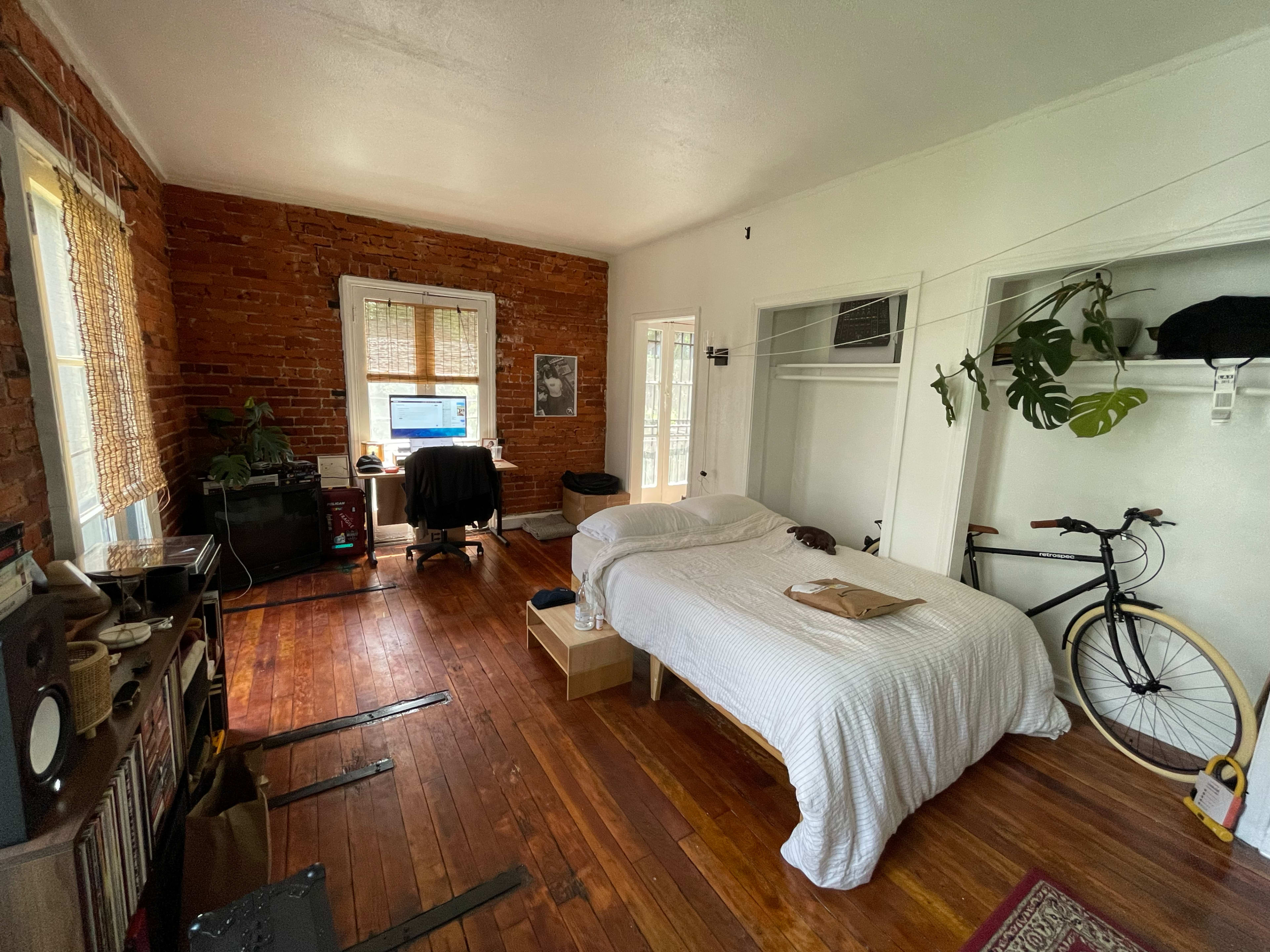 The image depicts a cozy bedroom with exposed brick walls, a bed covered with a white blanket, a desk with a computer, a bicycle leaning against the wall, and wooden flooring.