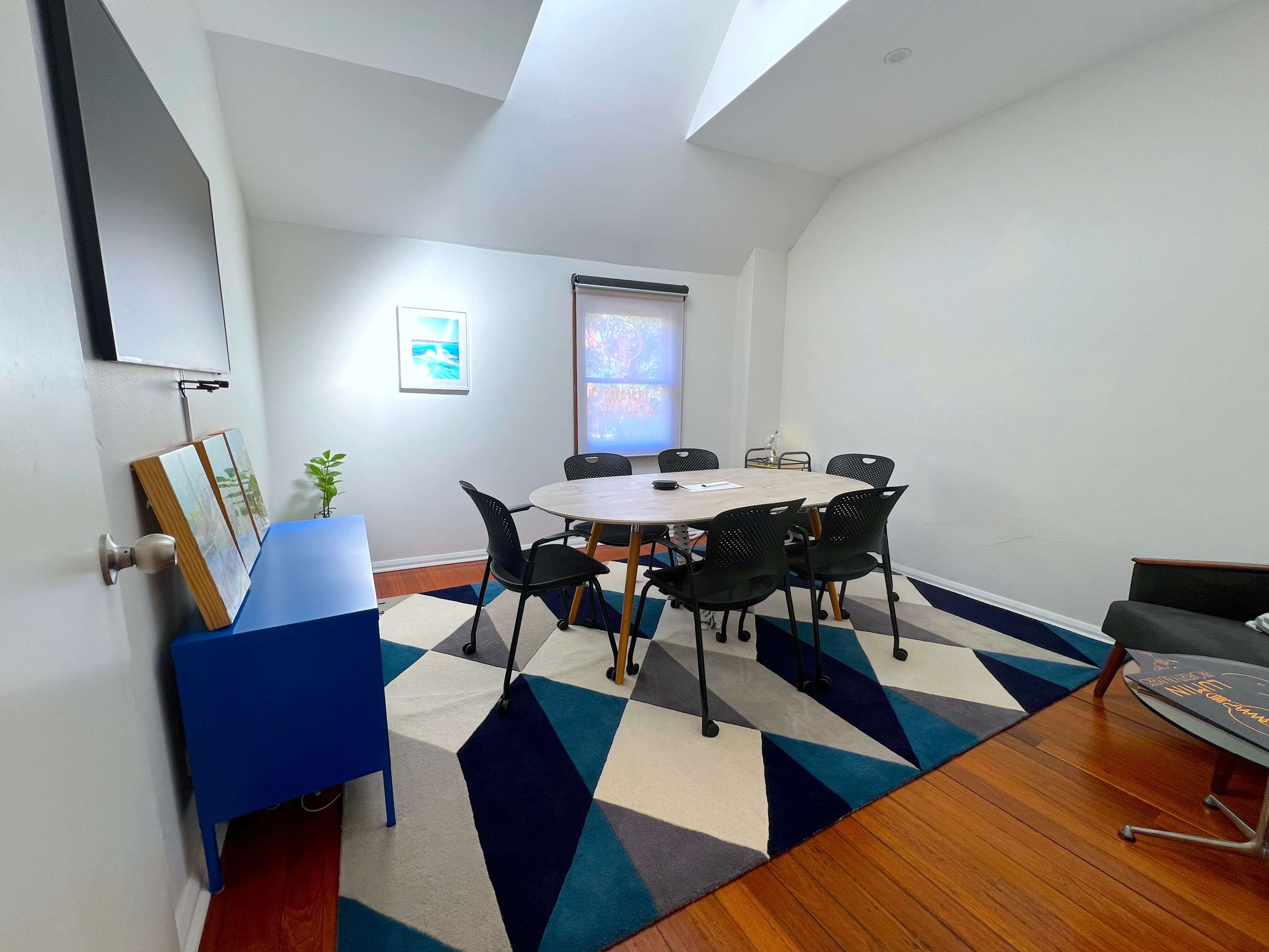 A conference room features a round table surrounded by black chairs on a patterned rug, with a television mounted on one wall and a plant on a blue sideboard.
