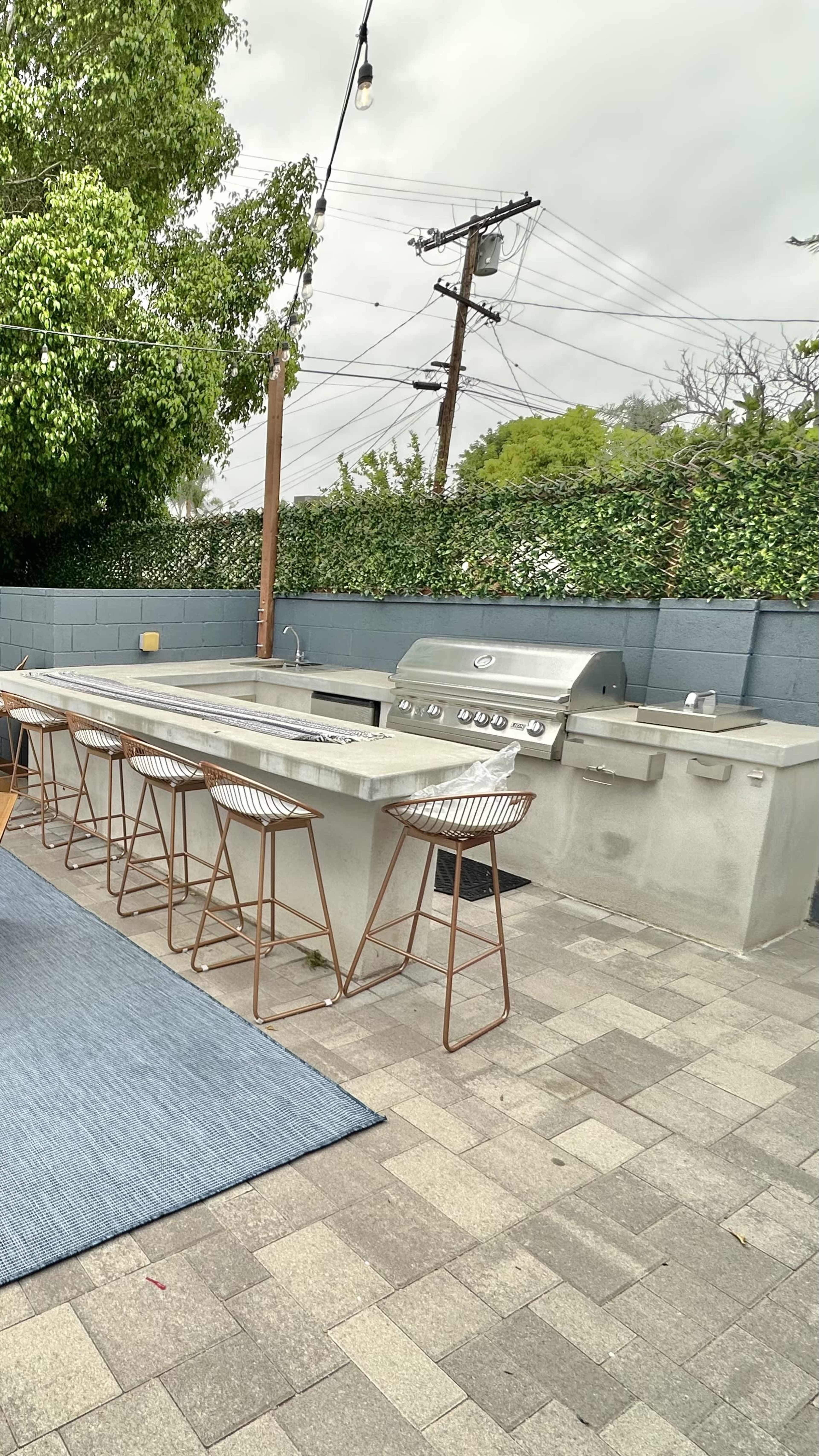 An outdoor kitchen with a stainless steel grill, a countertop, and bar stools under a cloudy sky.