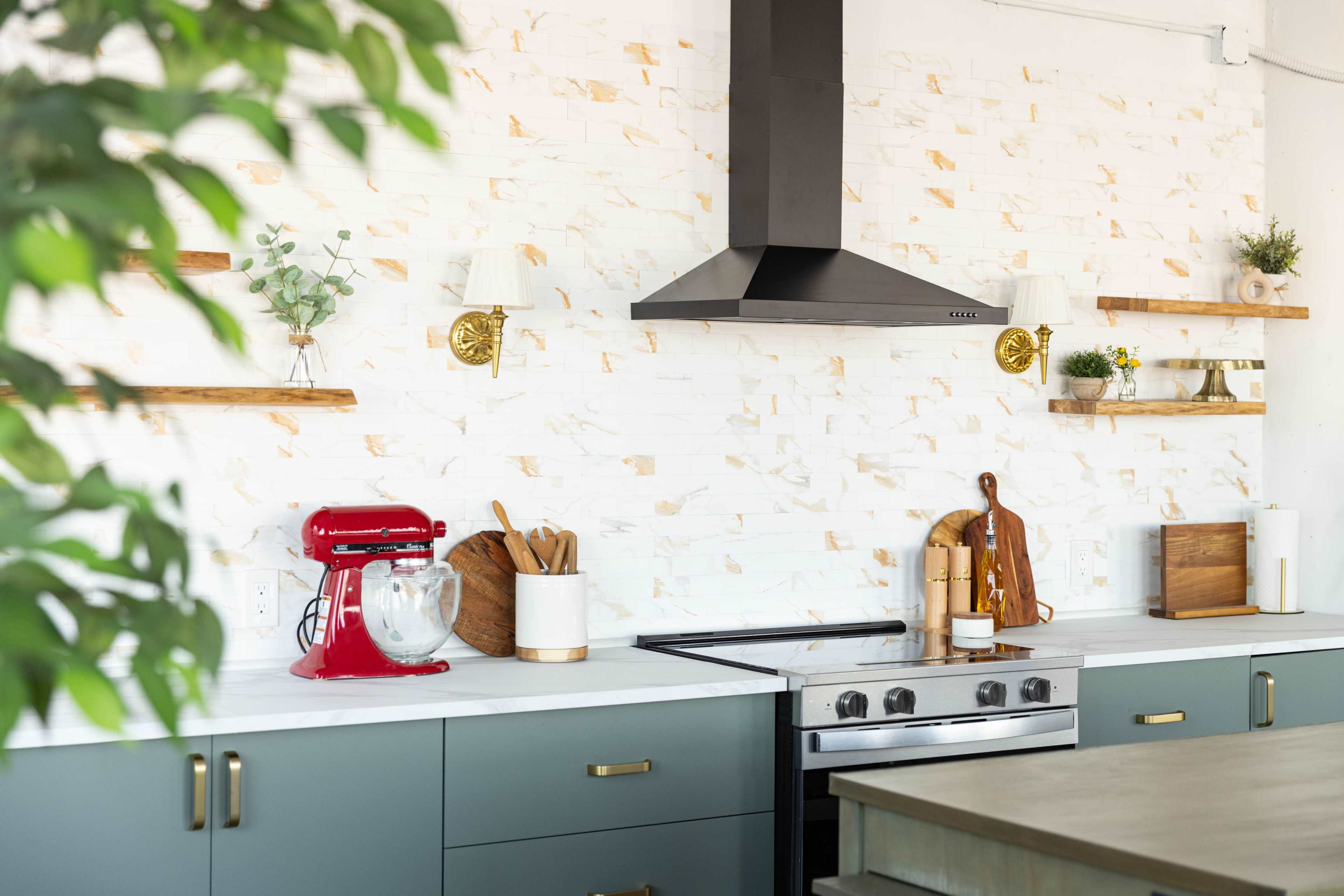 The image shows a modern kitchen featuring a mix of dark green cabinetry, a stainless steel stove, and a red stand mixer against a white brick wall with wooden shelves and decorative plants.