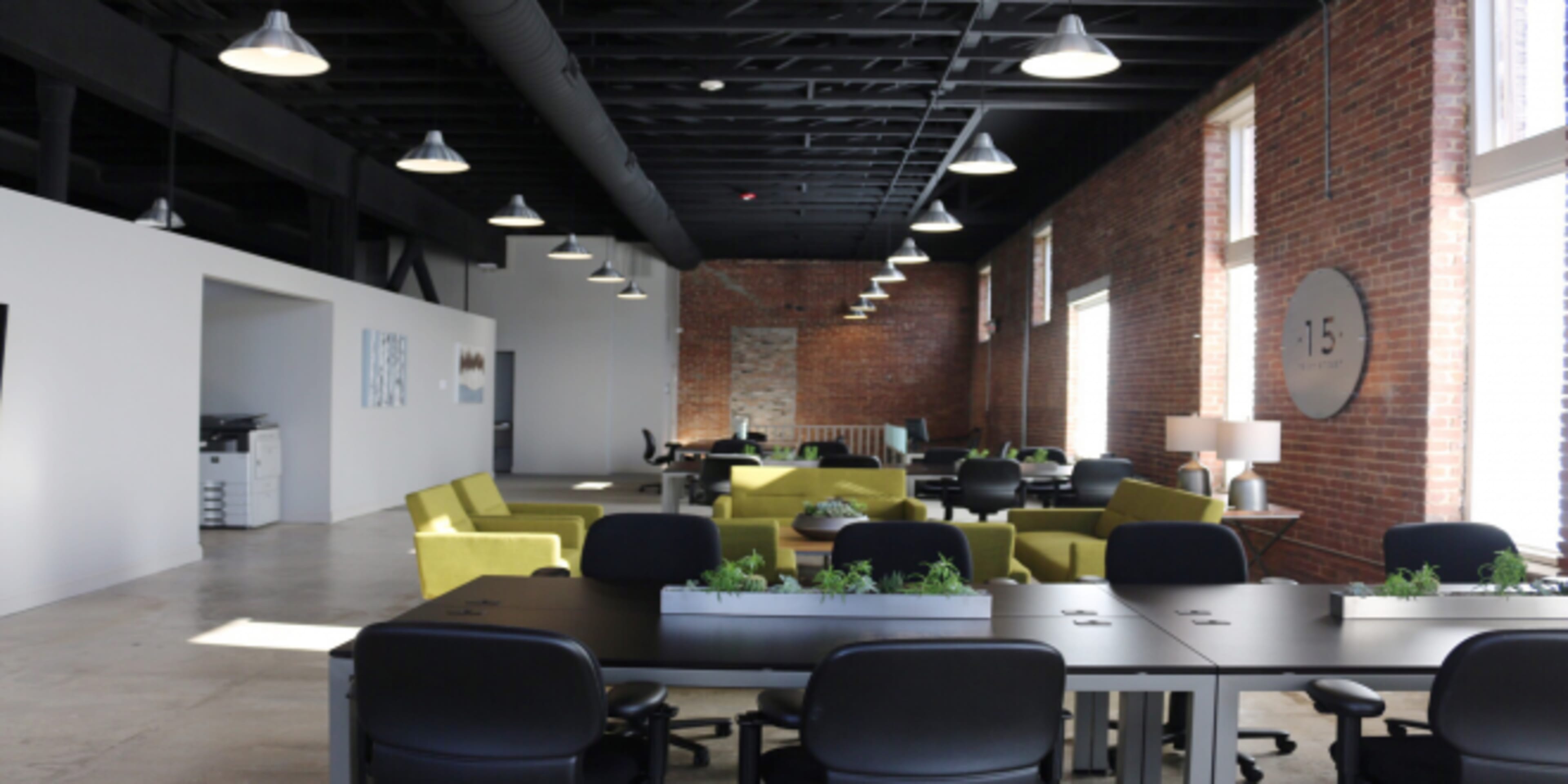 A modern office space with brick walls, long communal tables, and seating areas featuring green chairs, illuminated by pendant lights.