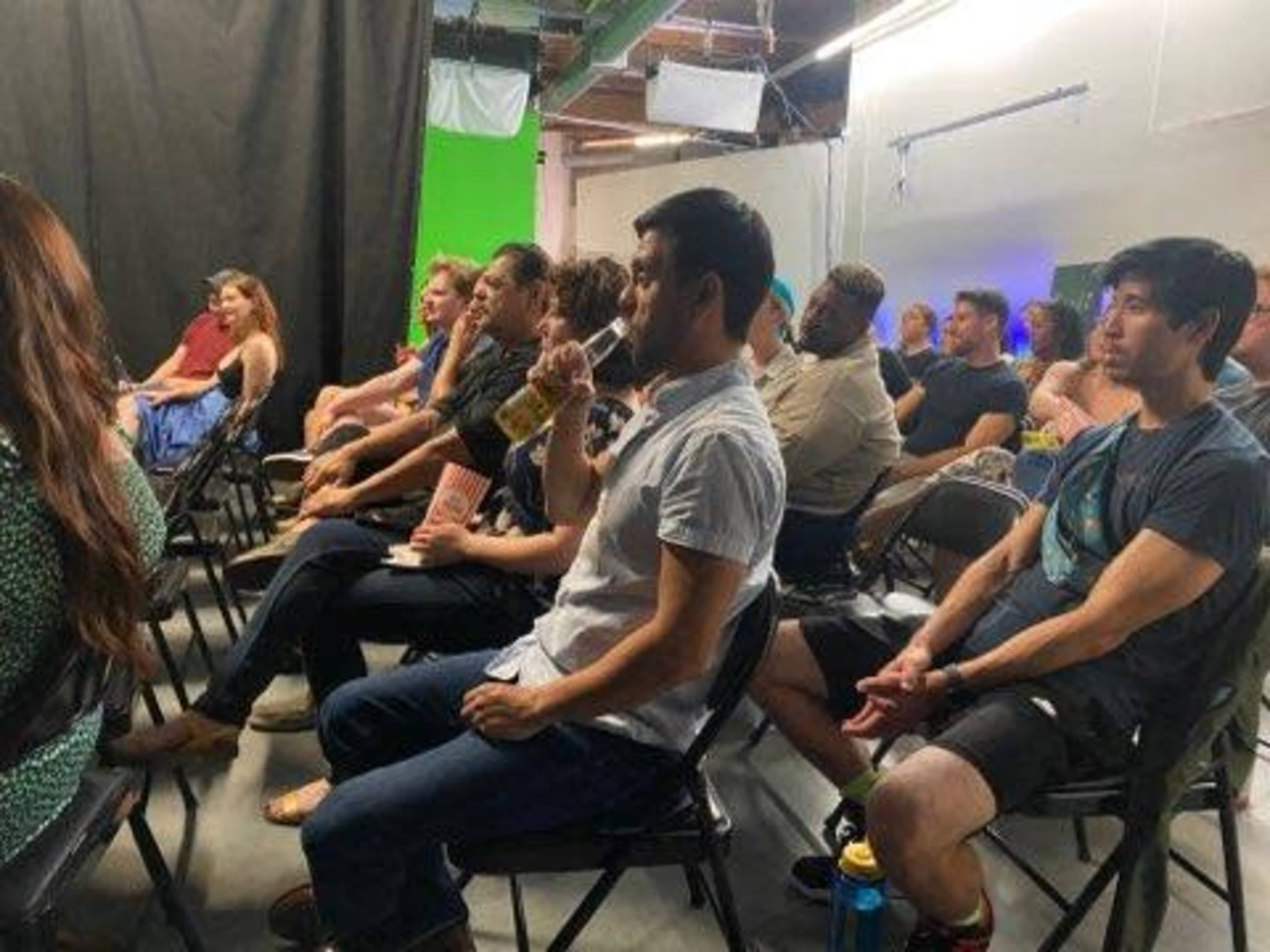 A group of people is seated in a classroom setting, facing the front, with a green screen backdrop.