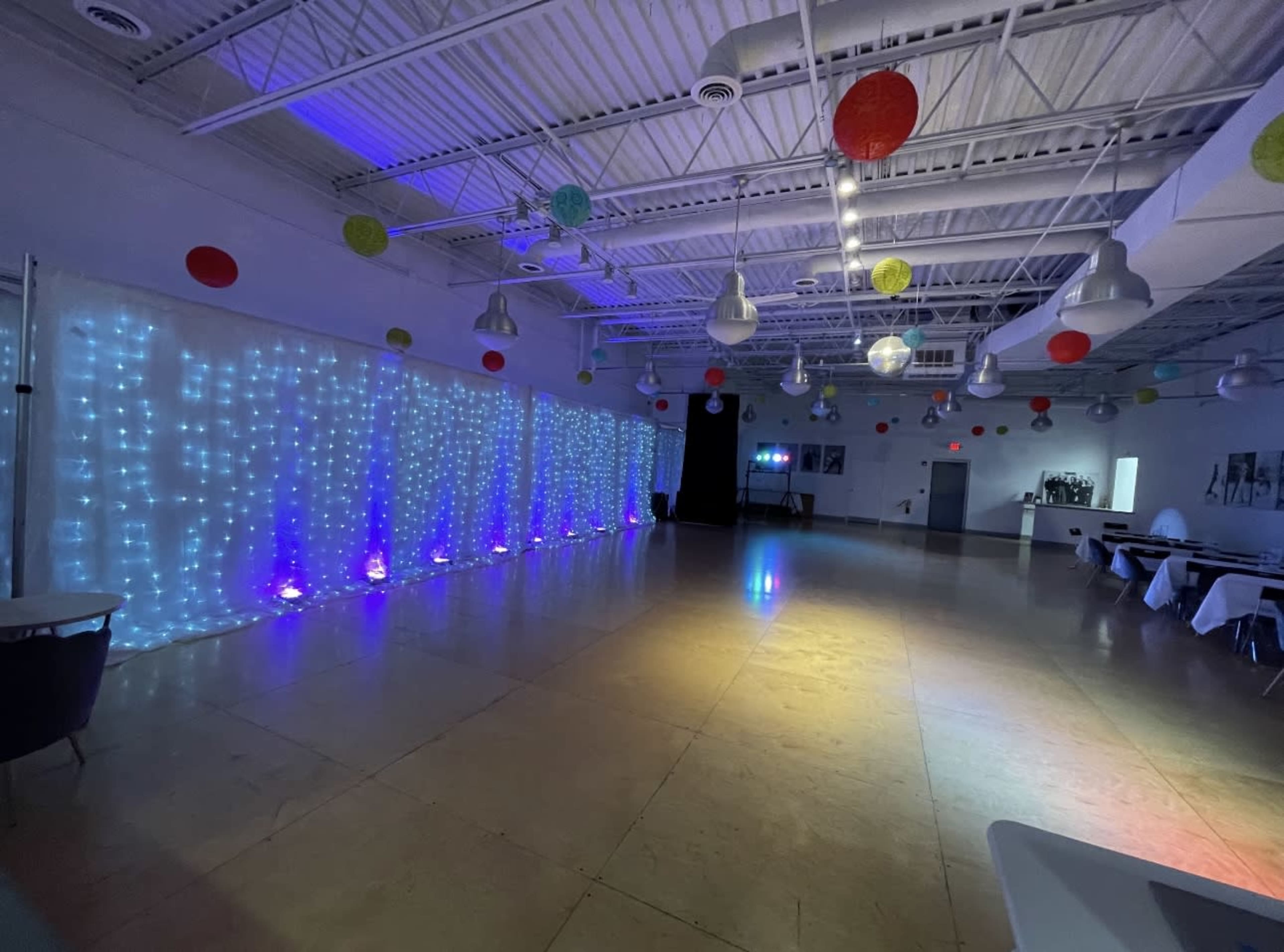The image shows a spacious event hall with illuminated walls featuring string lights, colorful decorations hanging from the ceiling, and tables set up along the sides.