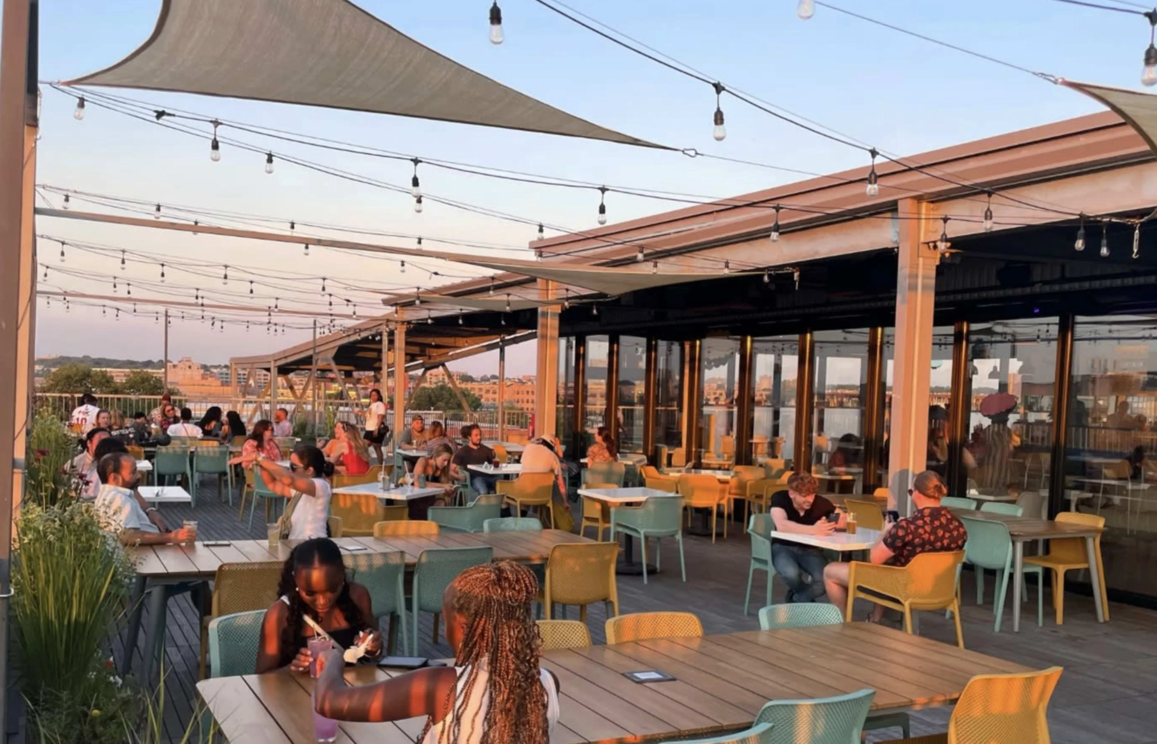 A rooftop restaurant with patrons dining at colorful tables under string lights during sunset.