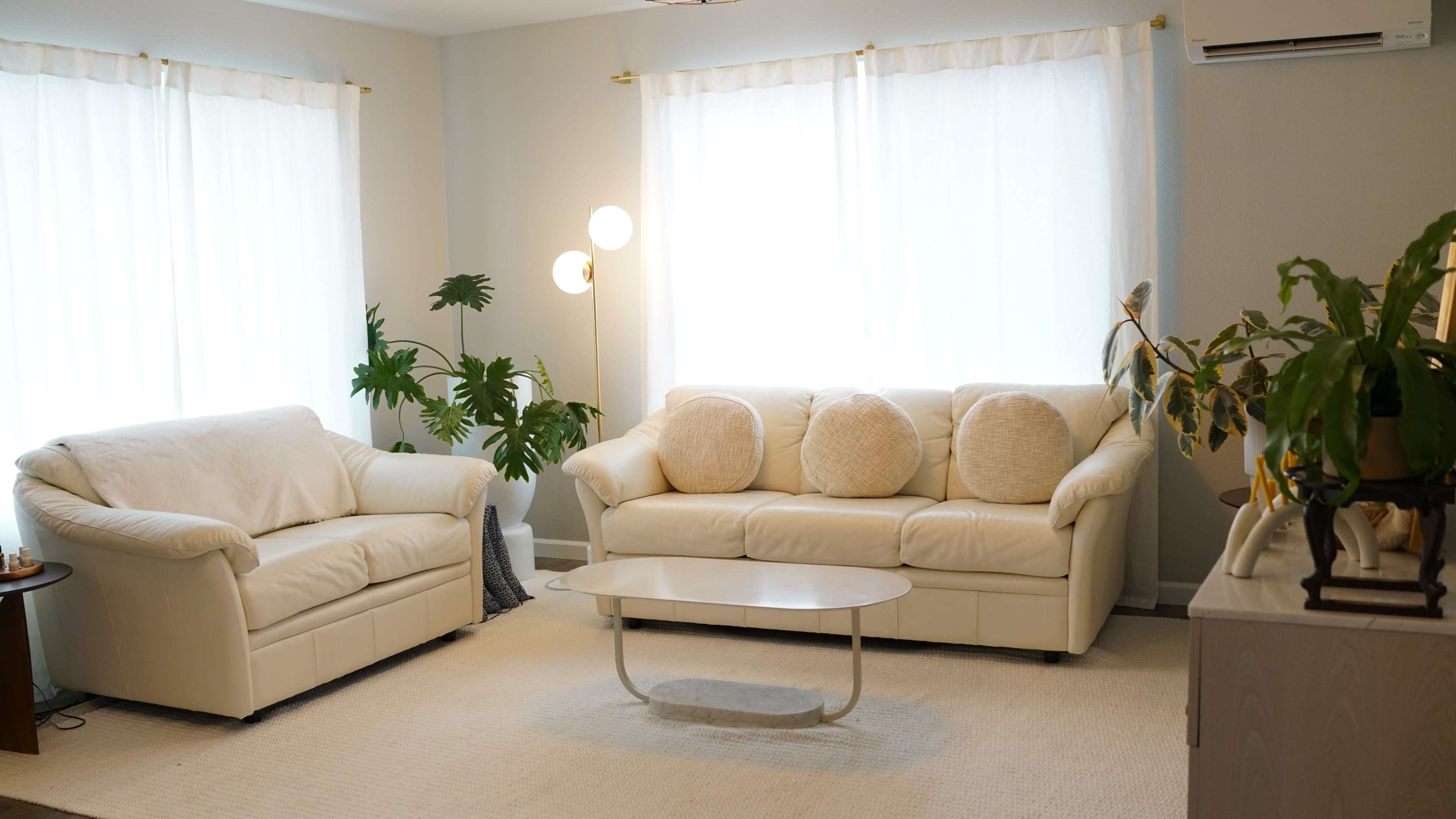A cozy living room features two white sofas, a glass coffee table, and large windows with sheer curtains, surrounded by indoor plants.