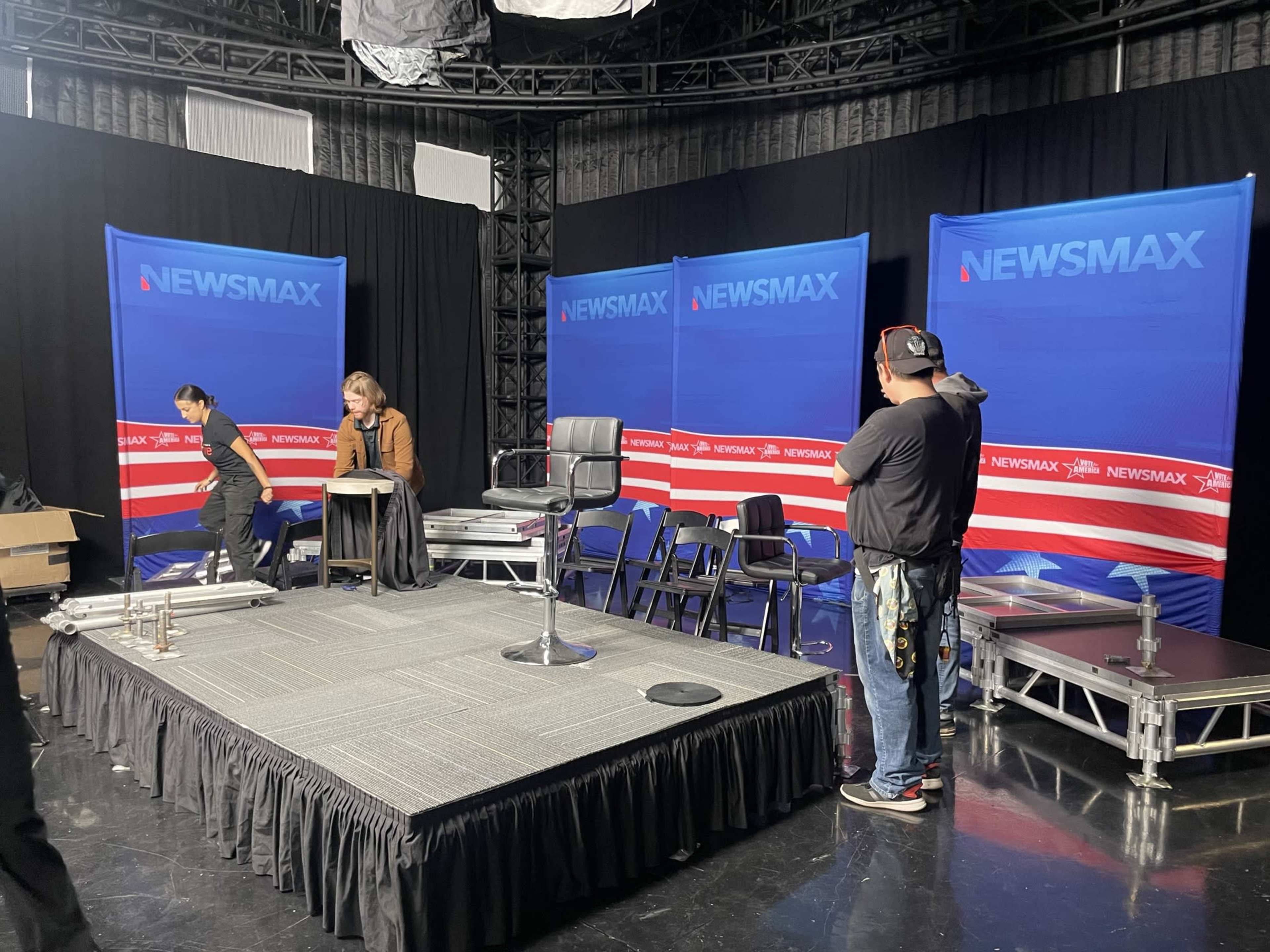 A television studio setup features a stage with a rotating chair and backdrop displaying the Newsmax logo, while crew members prepare the space.