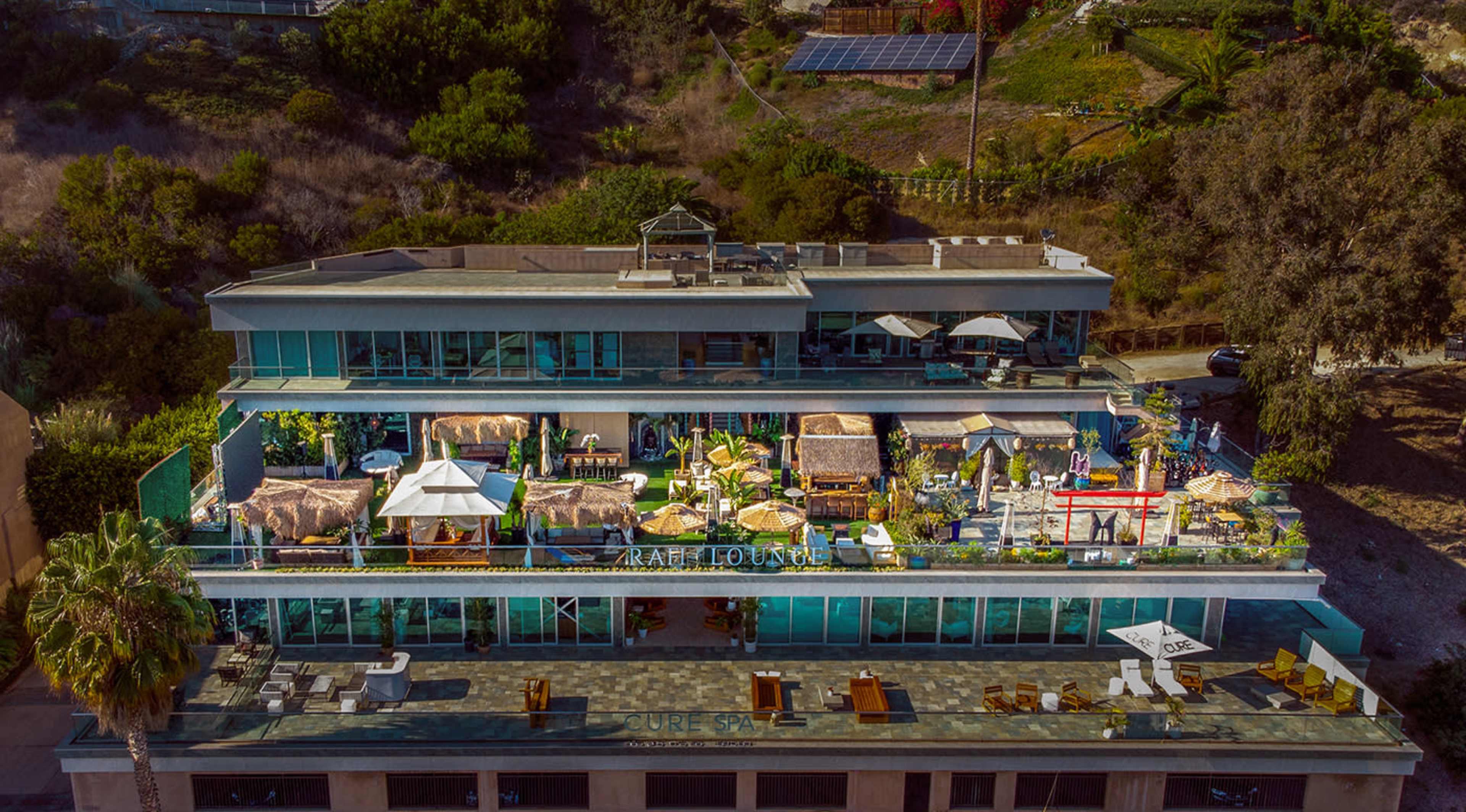 A multi-level building with a rooftop terrace featuring various lounge areas, seating, and decorative structures surrounded by greenery.