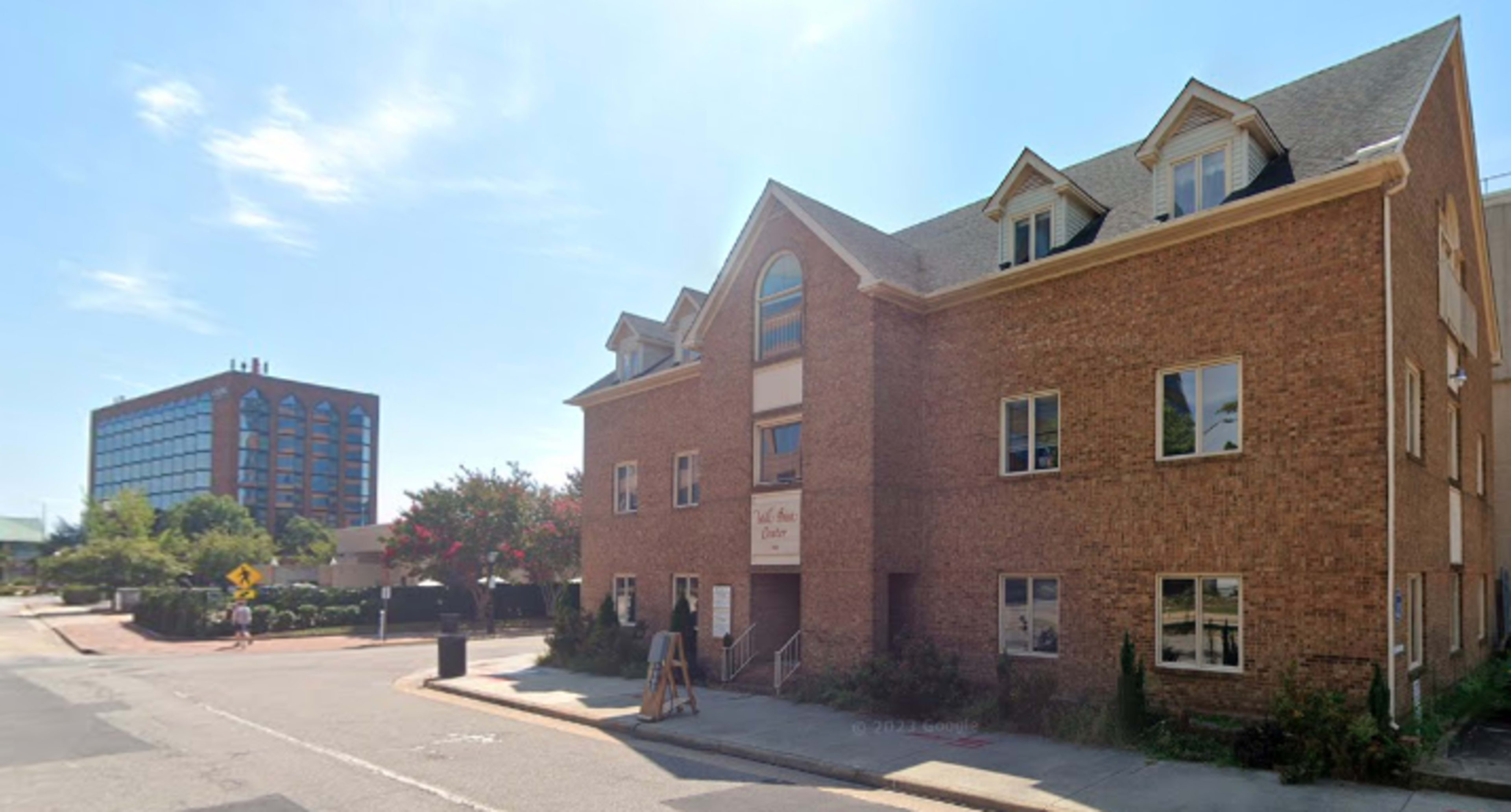 The image shows a three-story brick building with multiple windows, located on a corner near a glass-fronted office building.
