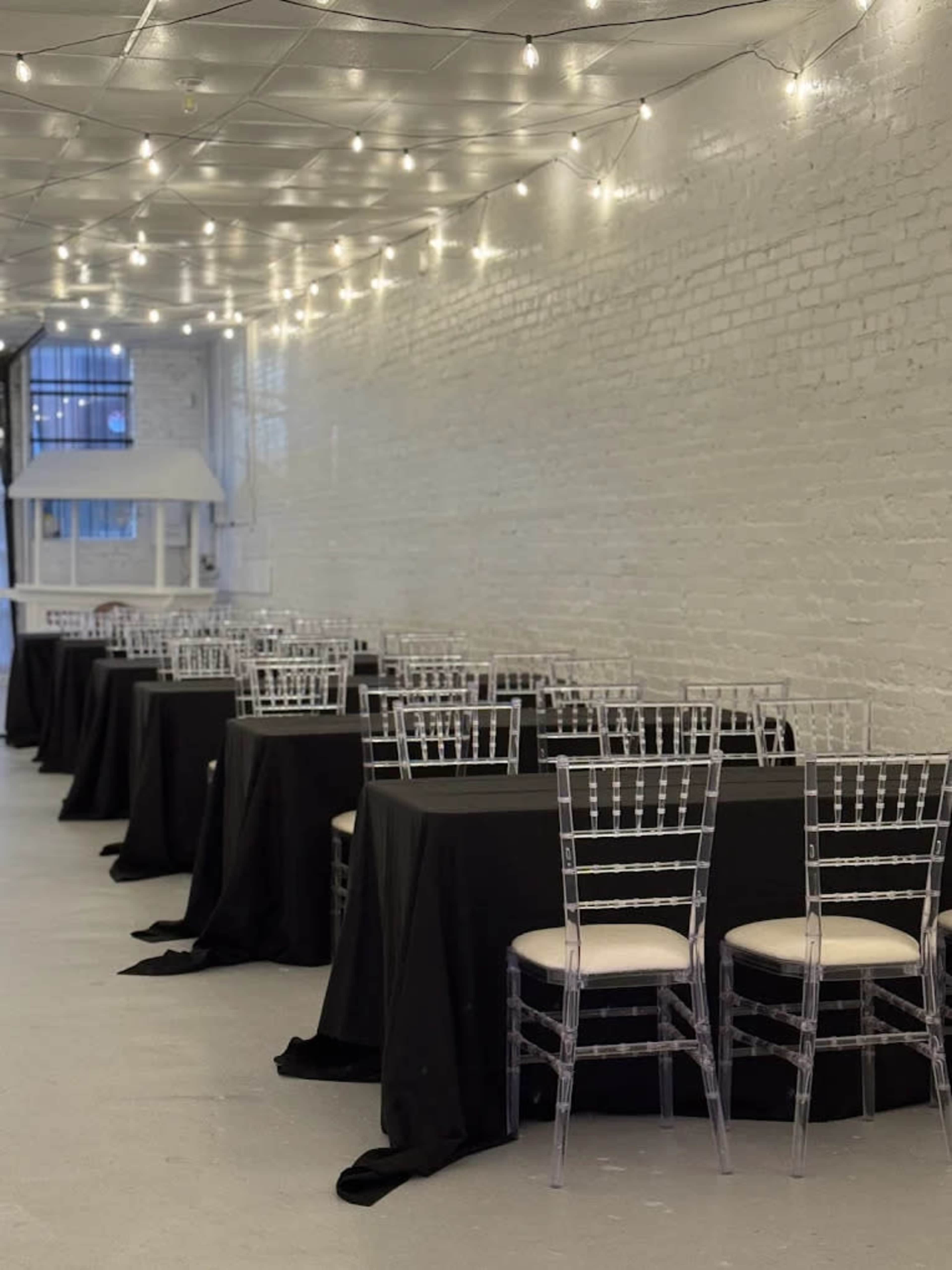 The image shows a long, brightly lit hall with several tables covered in black linens, each paired with clear acrylic chairs.