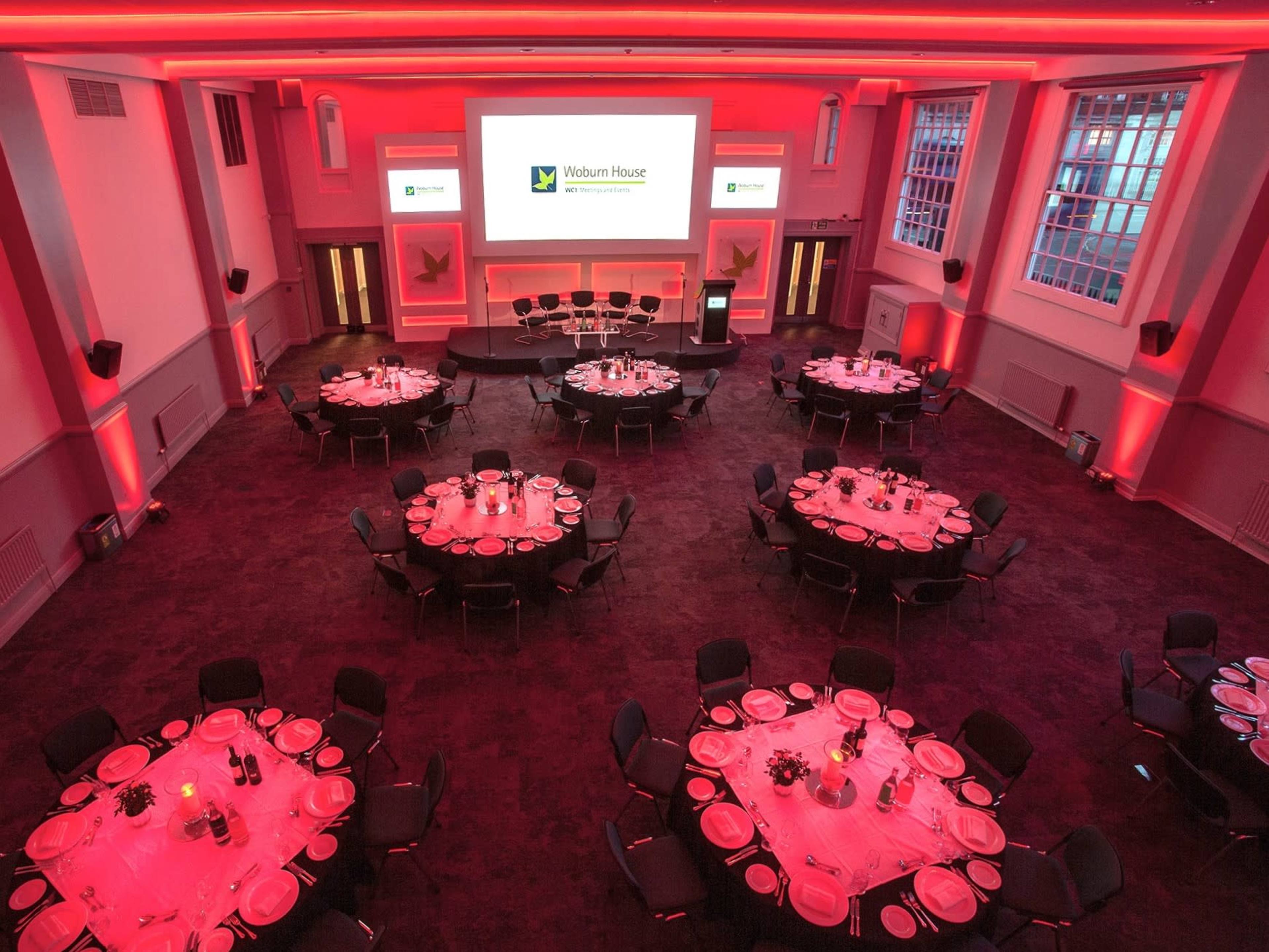 A banquet hall set up for an event, featuring several round tables with place settings, a stage with a screen, and red lighting illuminating the space.