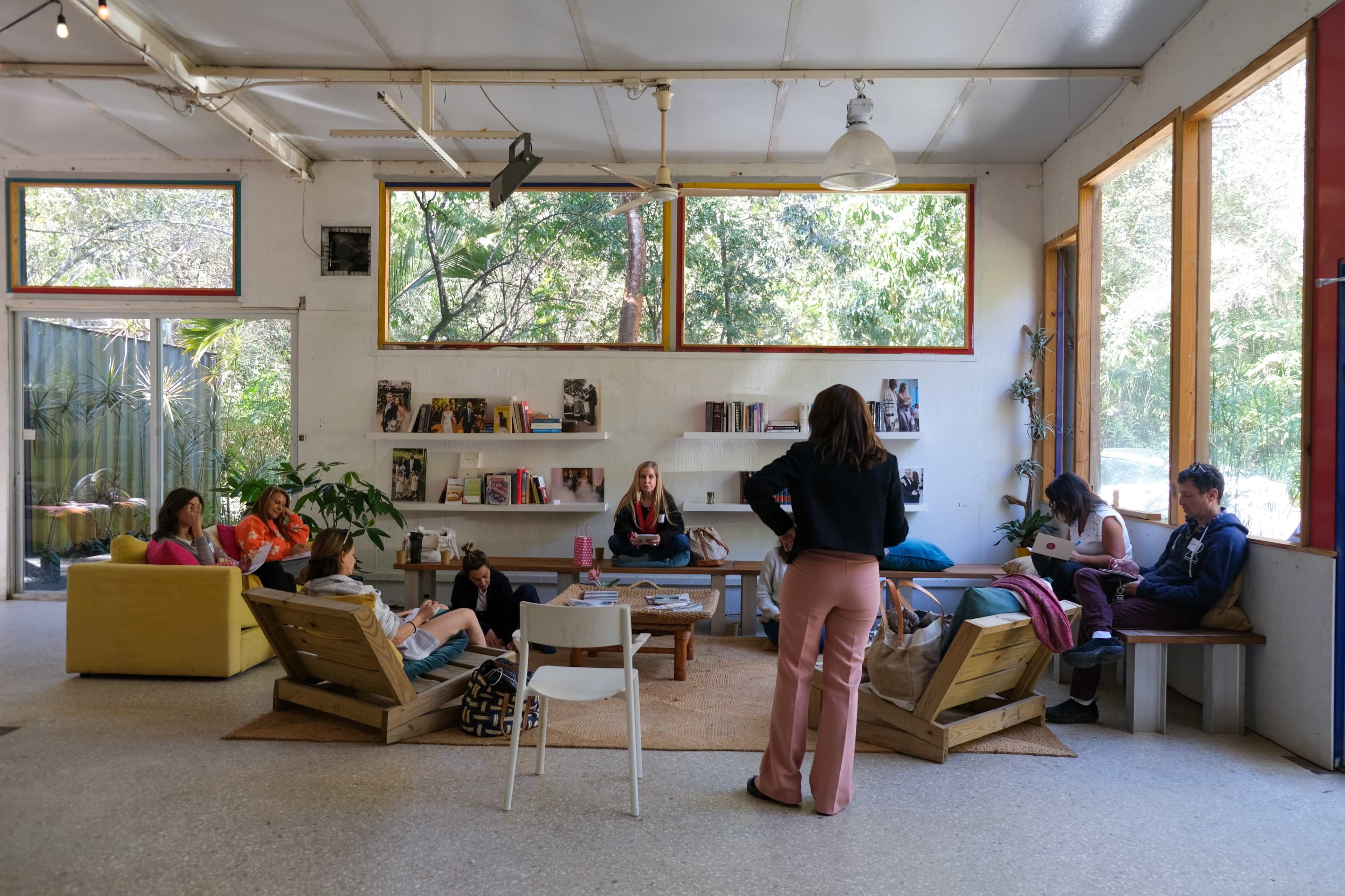 A bright, open space features a mix of seating arrangements and several people engaged in various activities, with large windows letting in natural light and greenery visible outside.