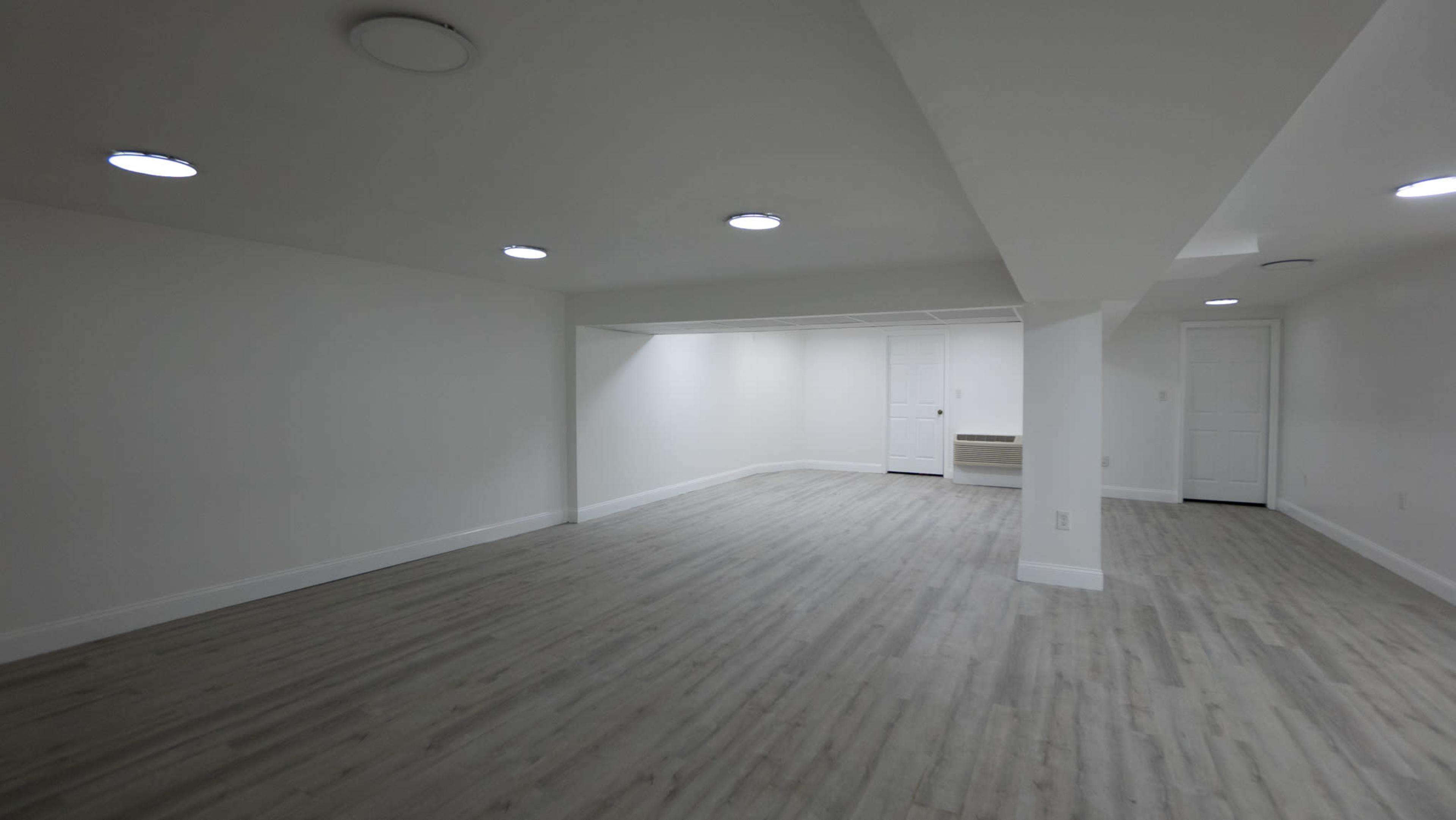 A spacious, empty basement room with light-colored walls and a pale wood-like floor, featuring recessed ceiling lights and double doors at one end.