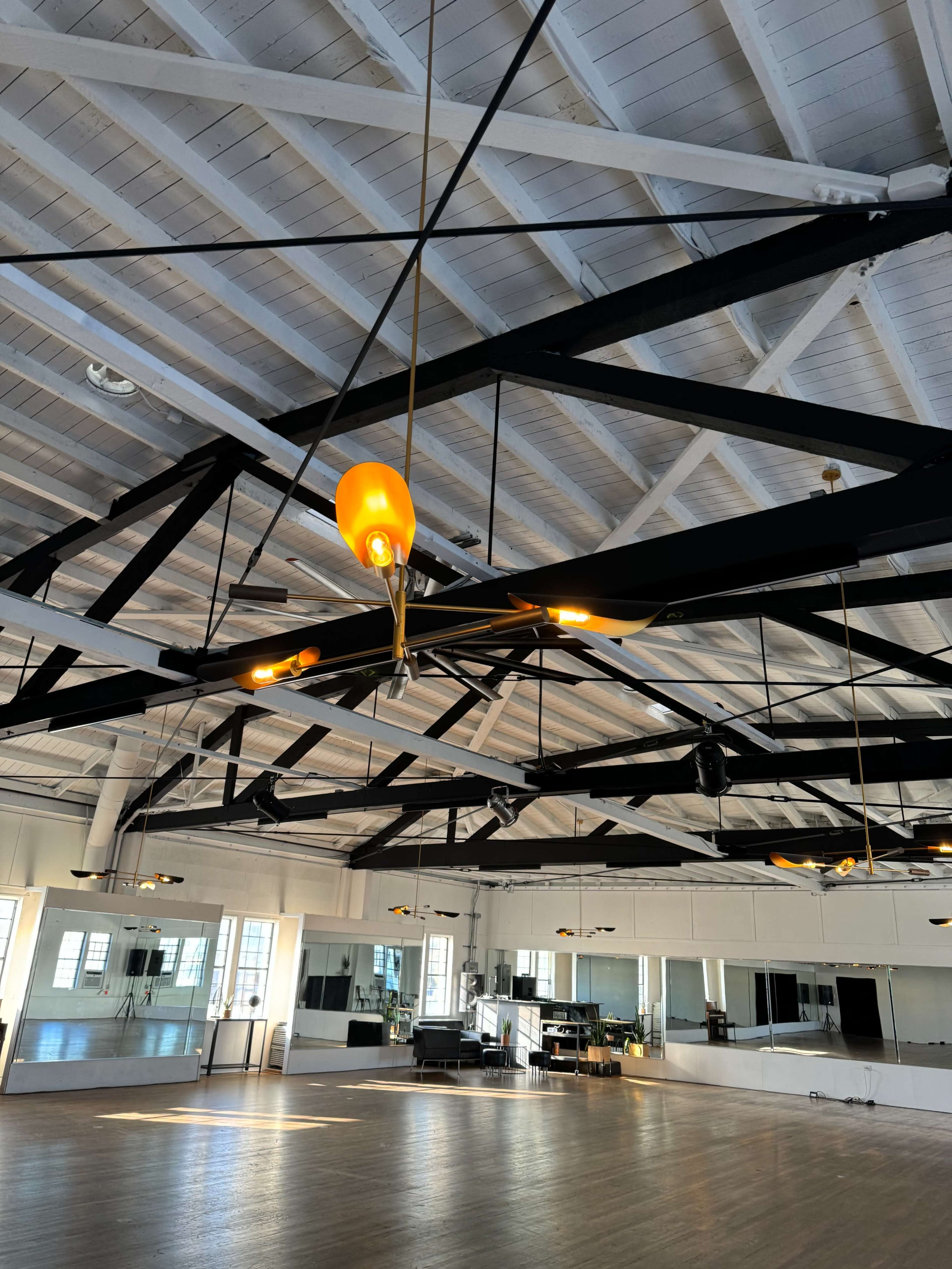 The image shows a spacious indoor dance studio with a wooden floor, large mirrors, and a ceiling featuring exposed beams and pendant lights.
