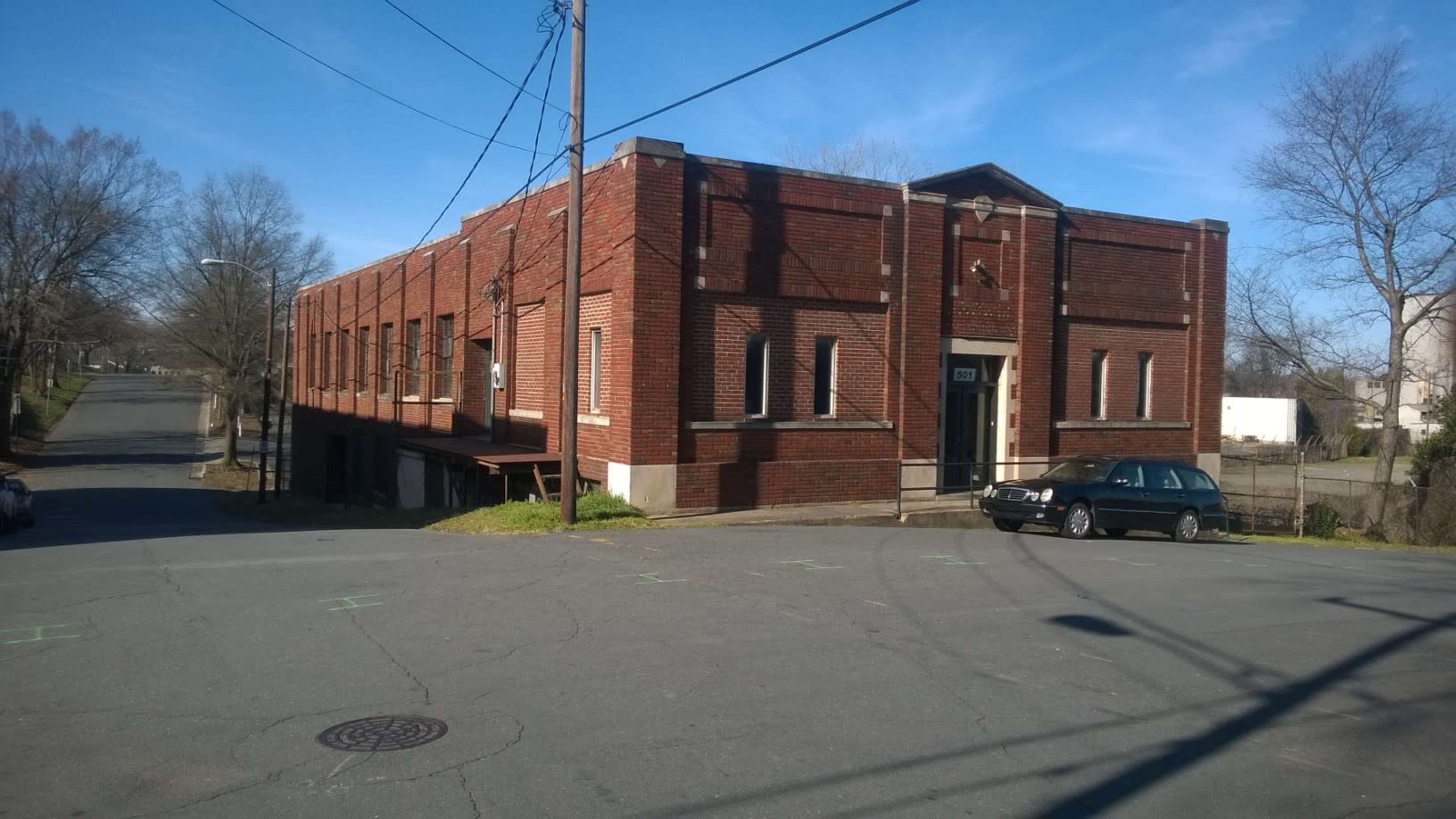 A red brick building sits at the corner of an intersection, with a parked car on the street and power lines overhead.