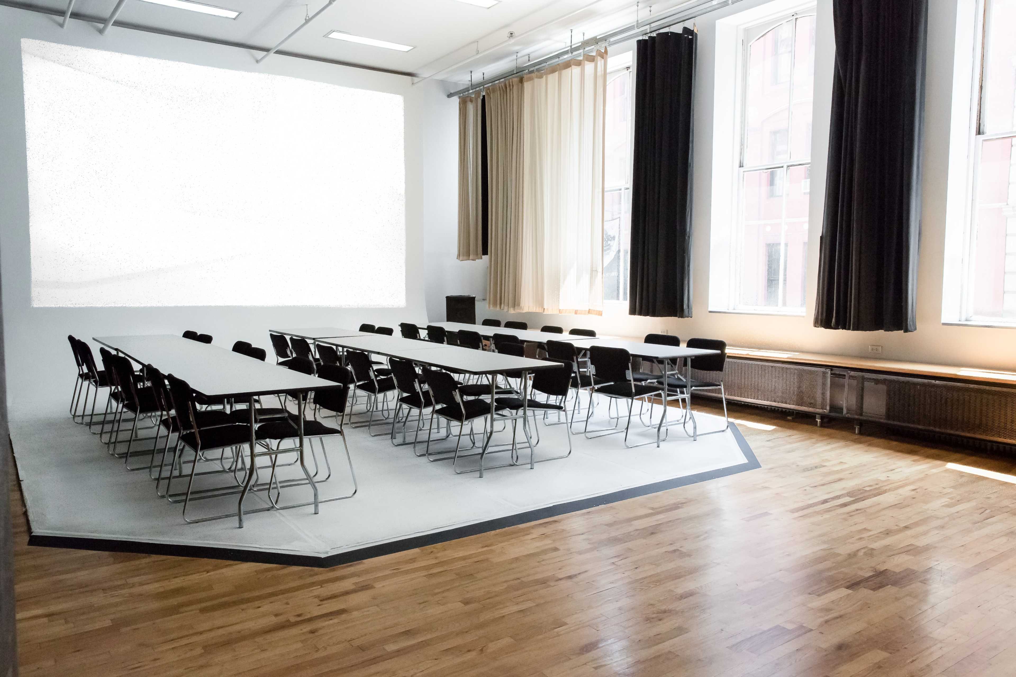 A large, empty conference room features a long table surrounded by black chairs, with natural light coming from tall windows and a blank wall for presentations.