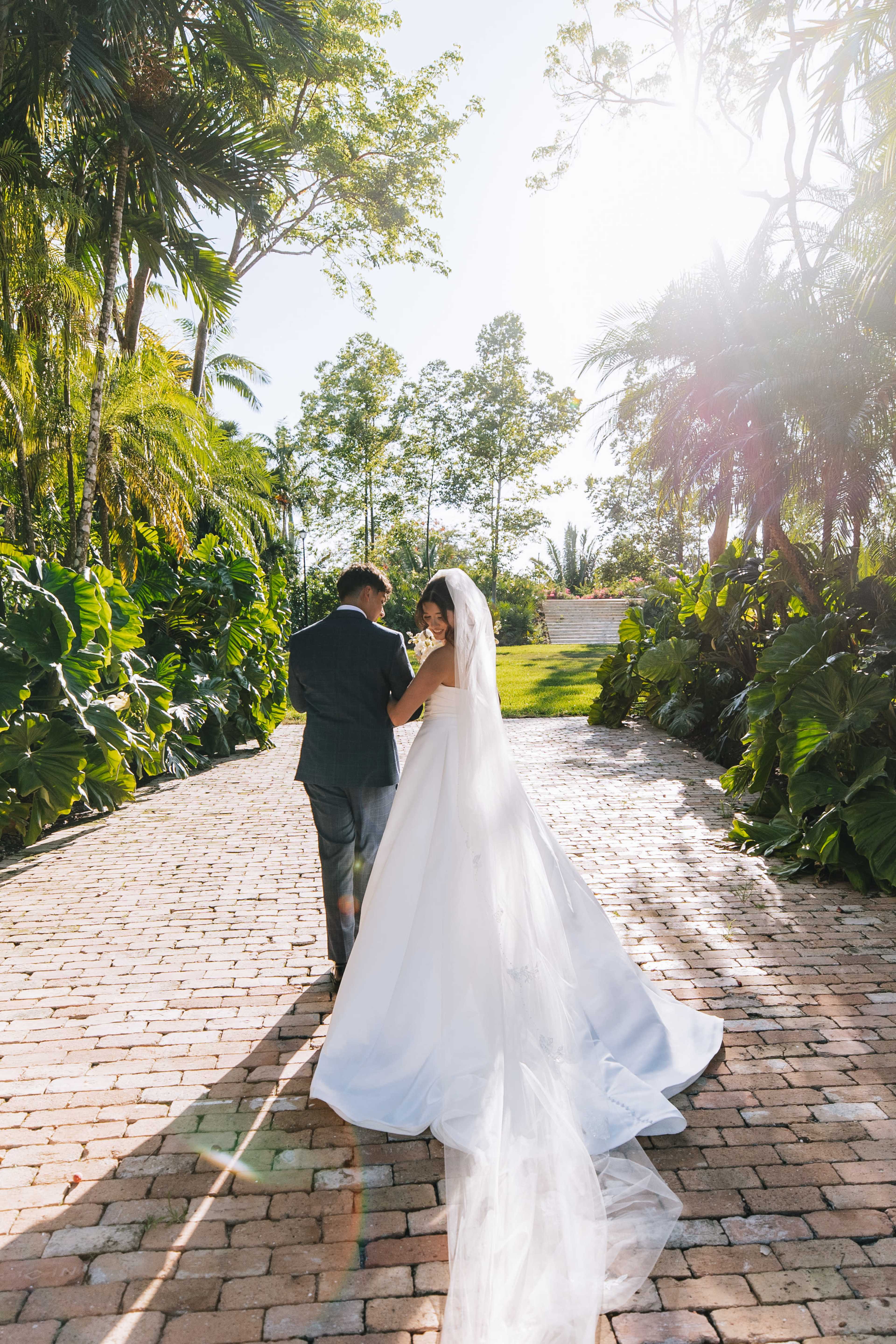 A couple walks hand in hand down a sunlit pathway surrounded by lush greenery, with the bride wearing a flowing white gown and veil.