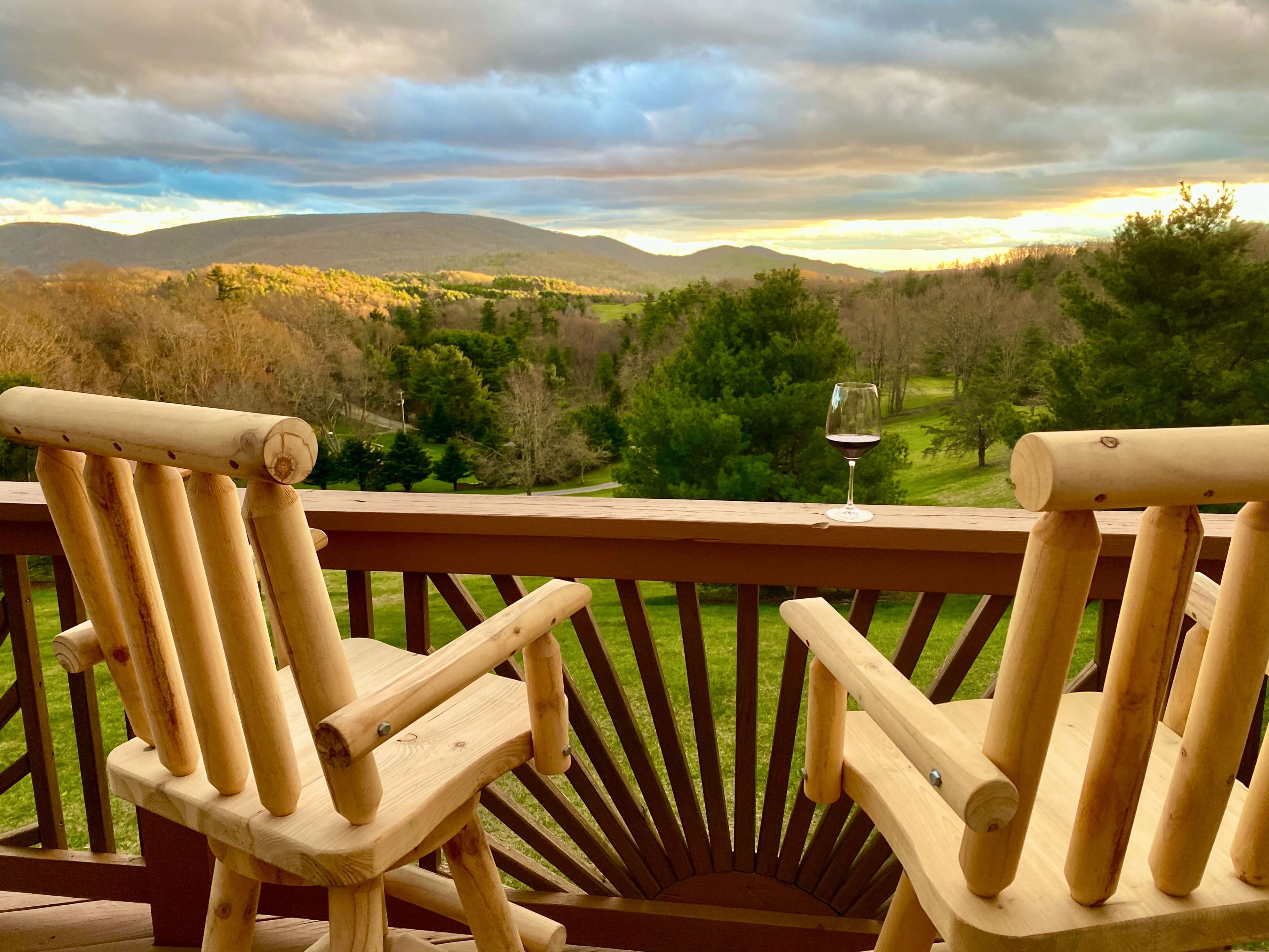Two wooden chairs are positioned on a balcony overlooking a landscape of hills and trees beneath a cloudy sky, with a glass of wine placed on the railing.