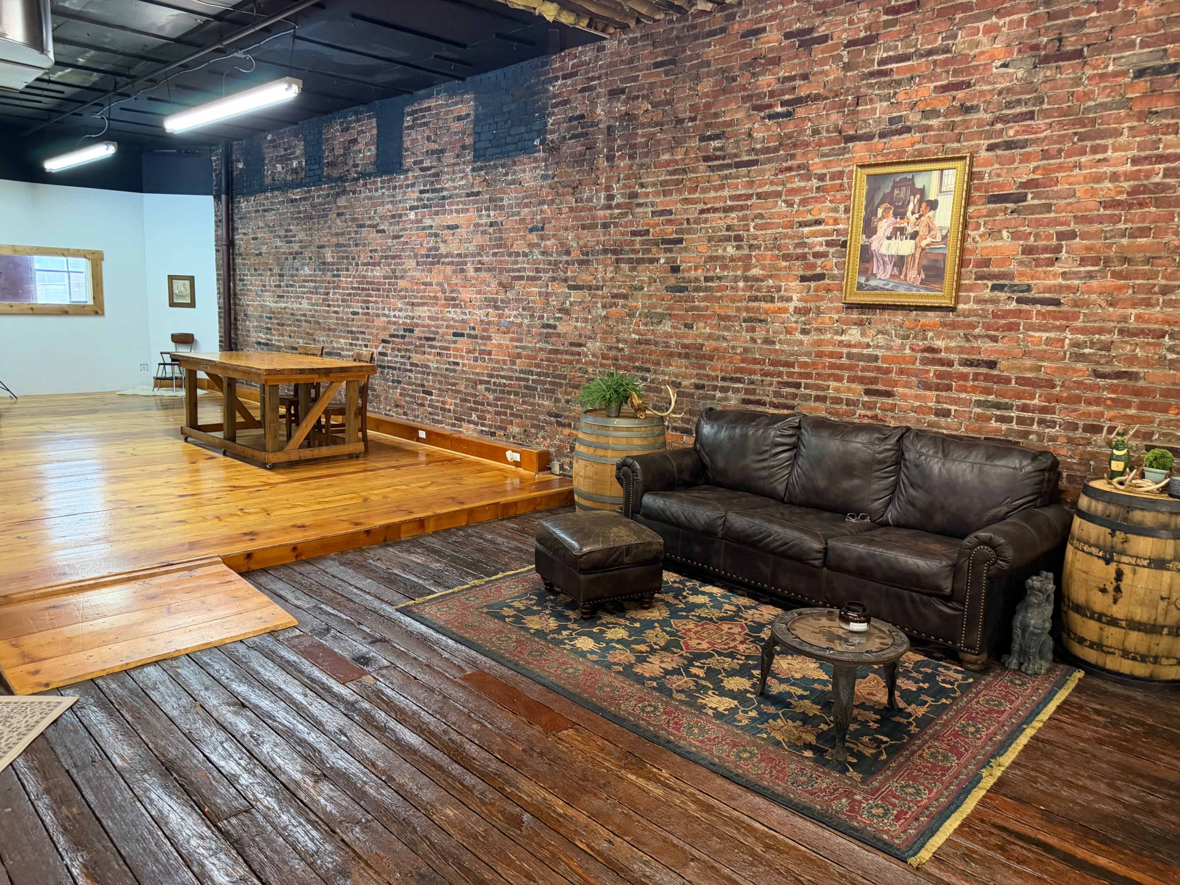The image shows a room with a brick wall, a wooden floor, a leather couch, a large table, and decorative barrels.