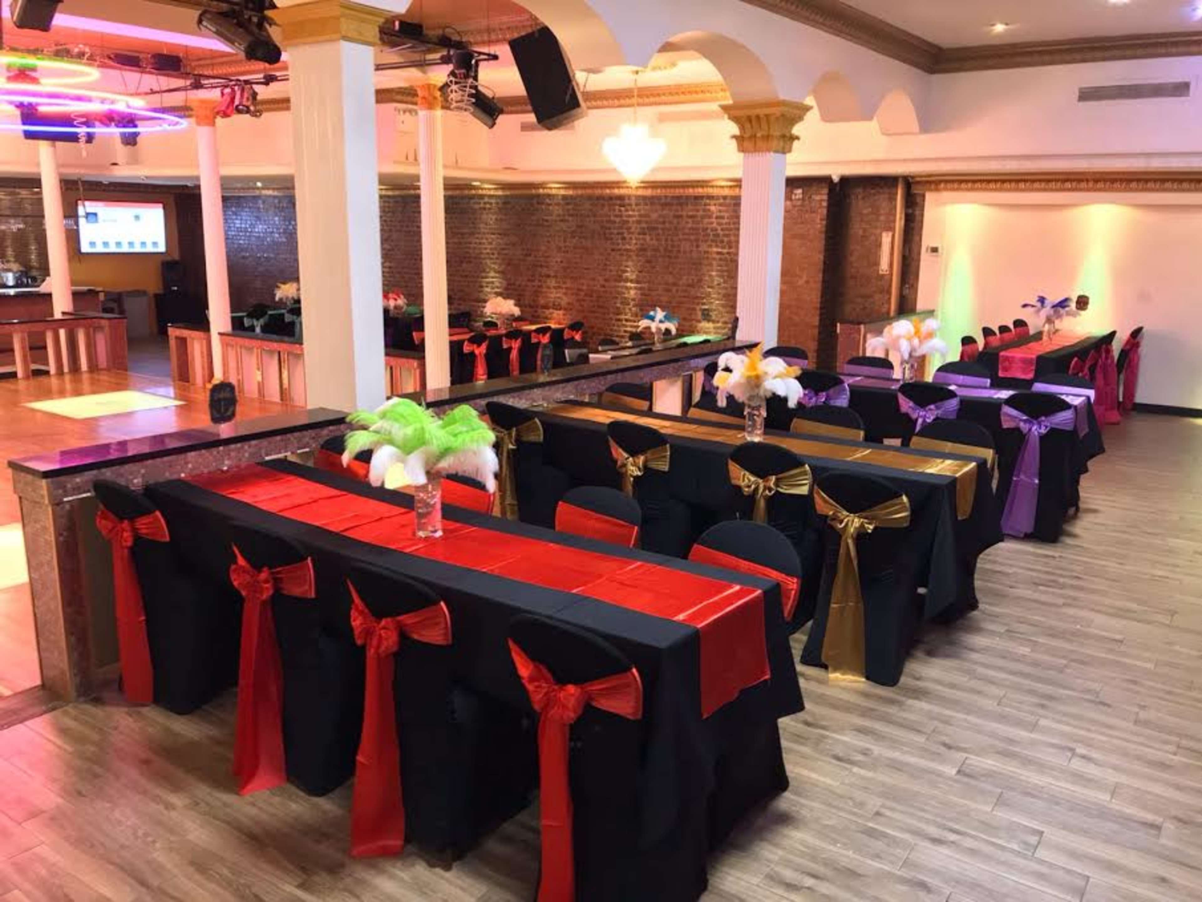 A banquet hall arranged with rows of tables covered in black cloth, decorated with colorful sashes in red, gold, and purple, and featuring centerpieces with feathers.