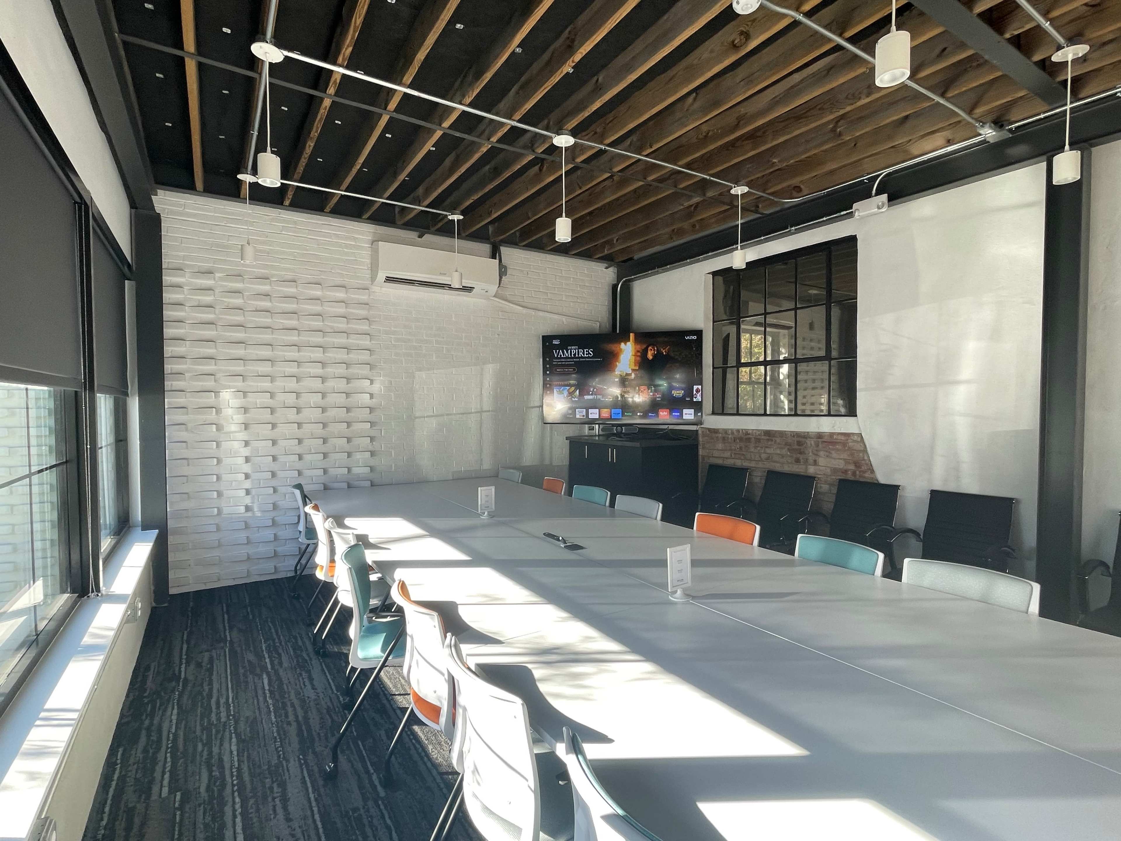 A modern conference room with a long table surrounded by chairs, featuring a large screen on one wall and windows allowing natural light to enter.