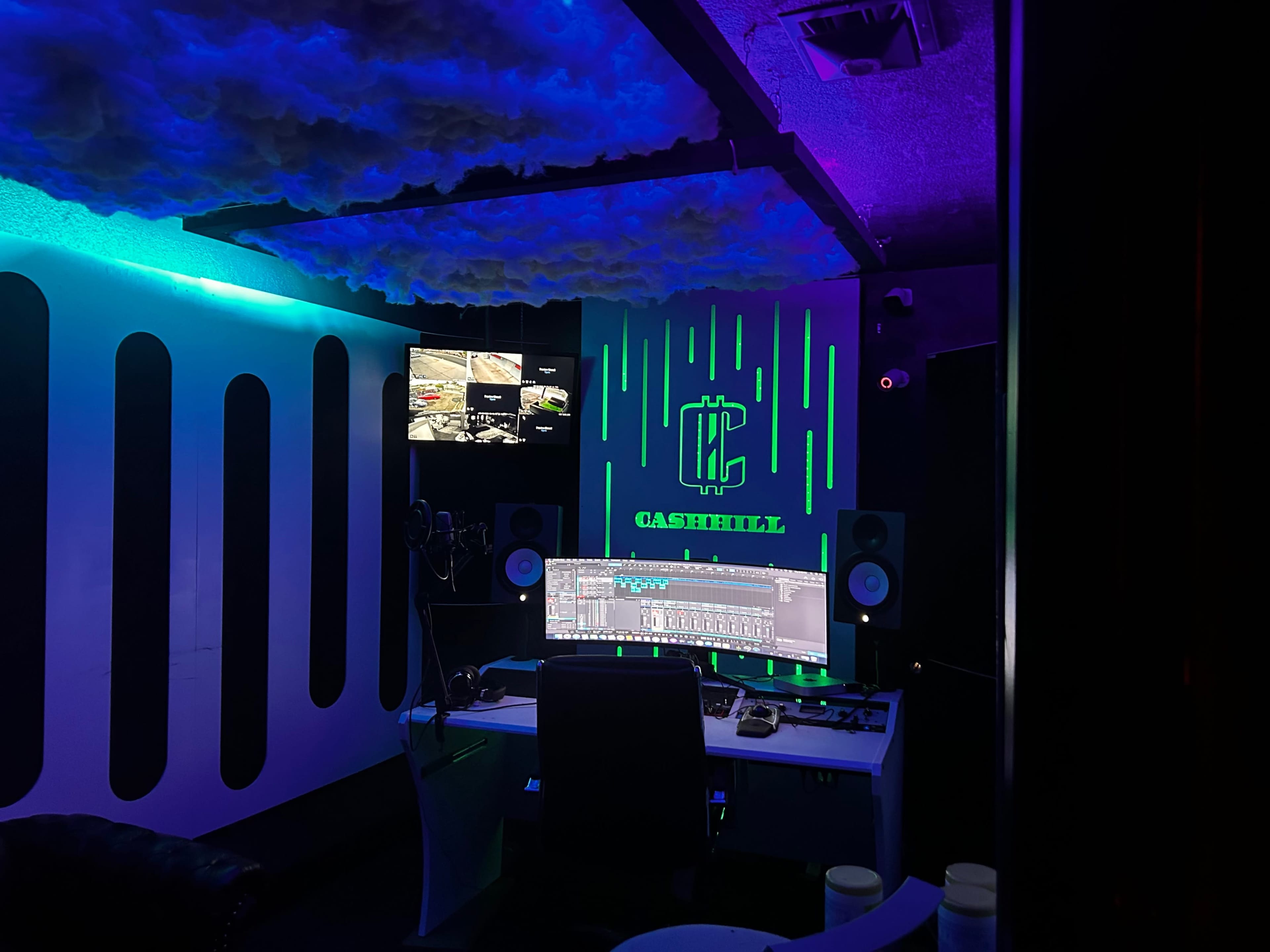 A modern recording studio with colorful lighting, a large computer monitor displaying audio software, and a sound booth on the side.