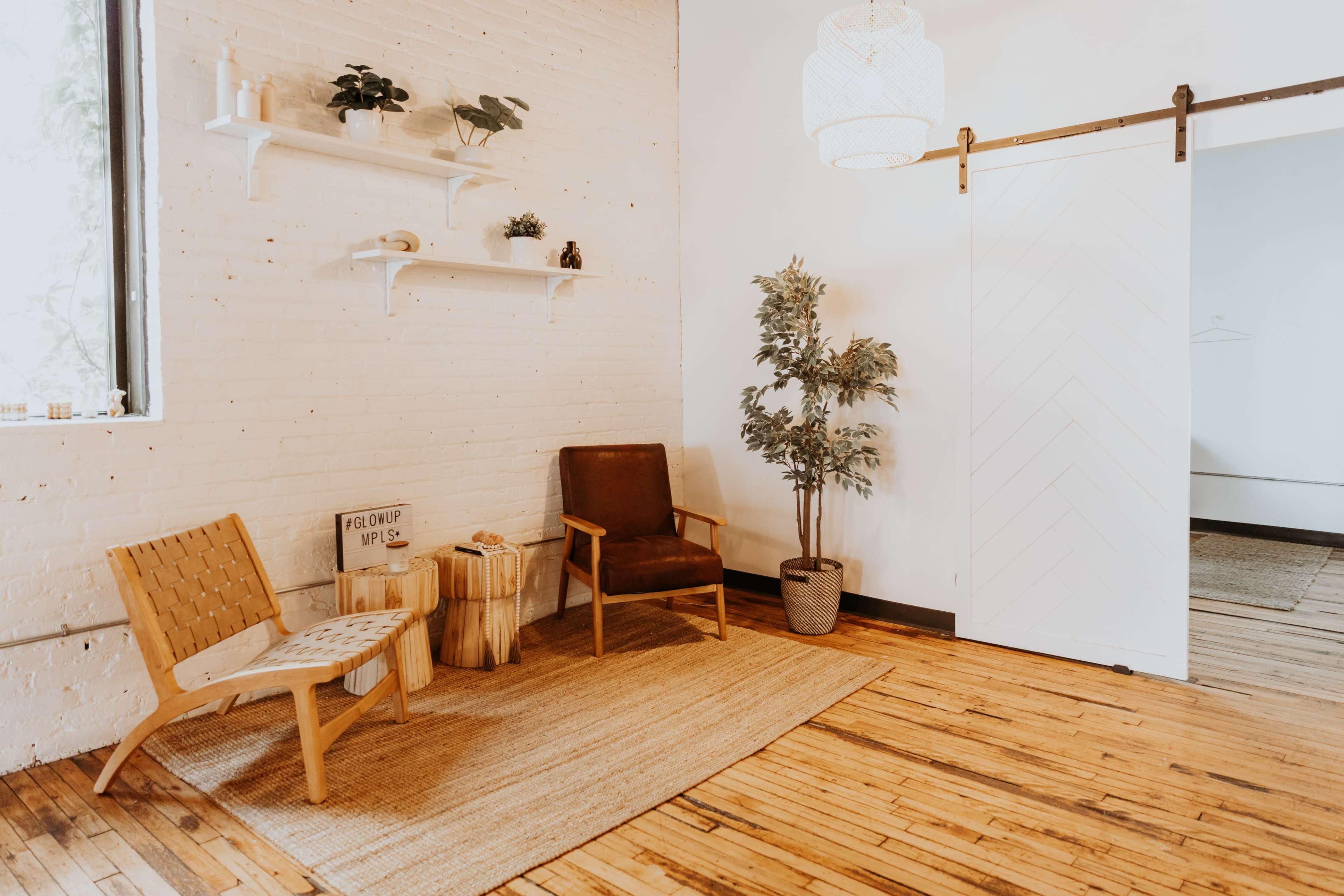 A light-filled indoor space with a wooden floor, featuring two chairs, a small side table, a potted plant, and a sliding door.