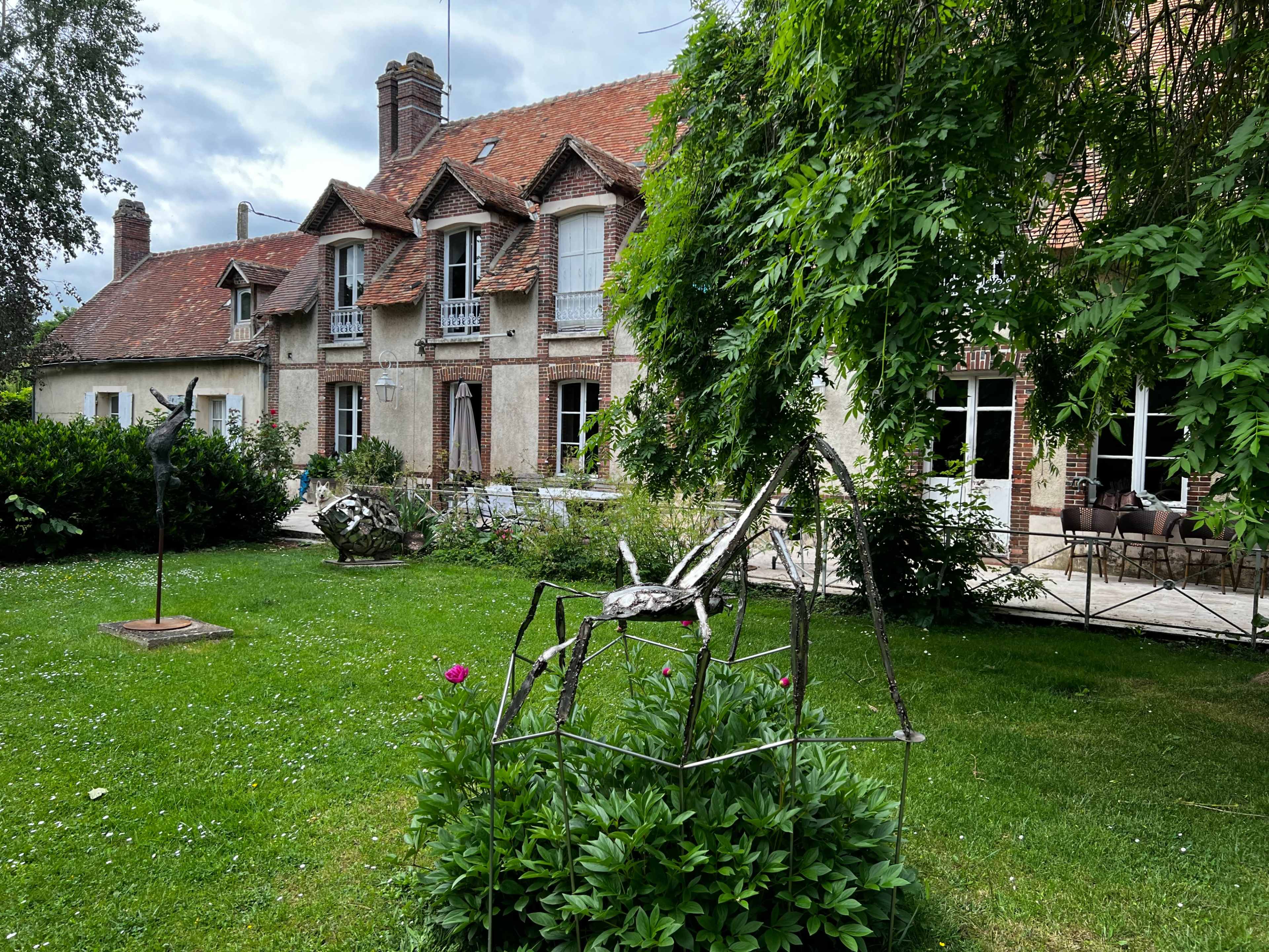 A large spider sculpture made of metal is positioned in a green garden in front of a two-story stone house with multiple windows and a sloping red roof.