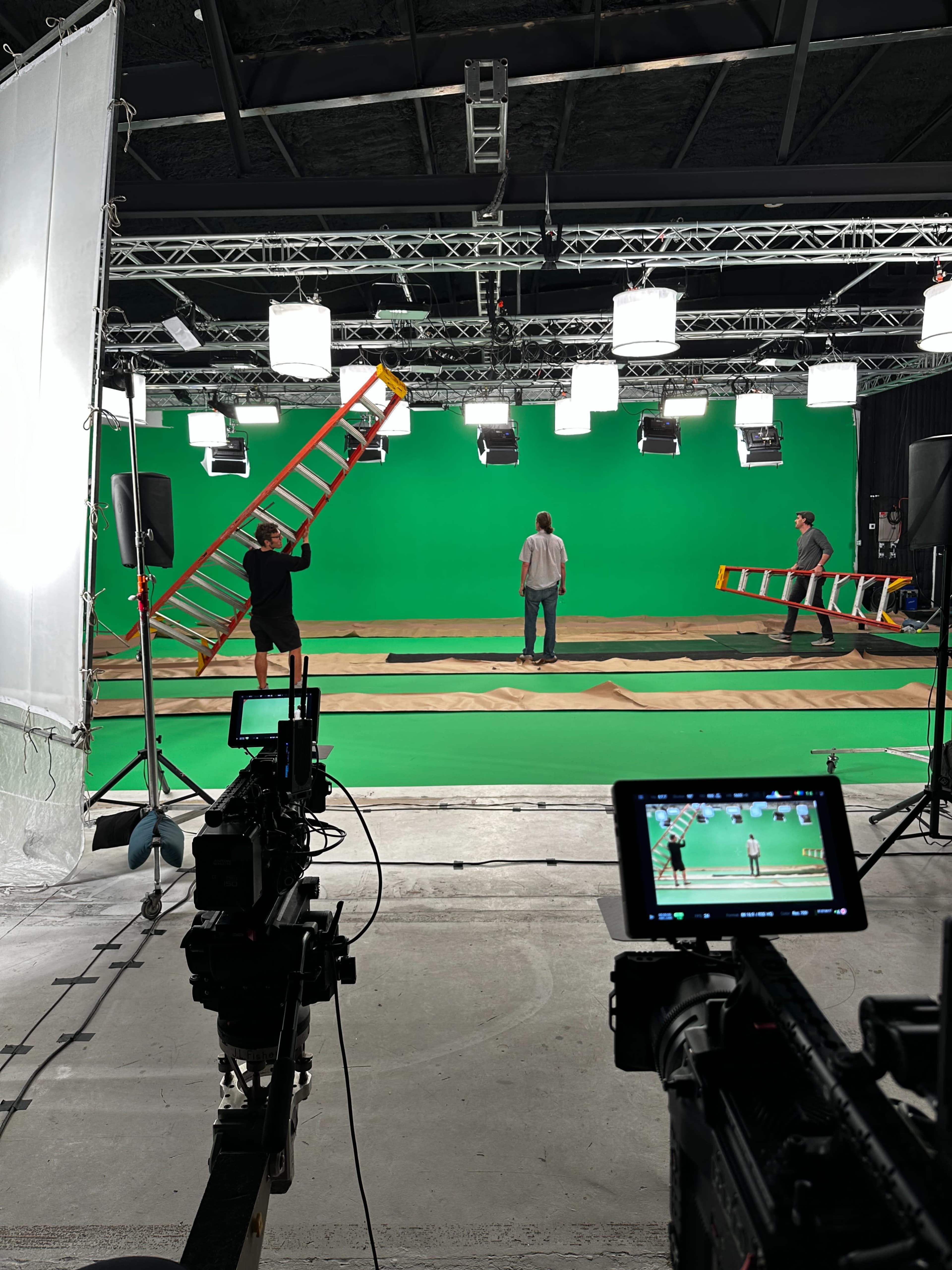 A video production studio is set up with a green screen backdrop, lighting equipment, and crew members preparing the scene.