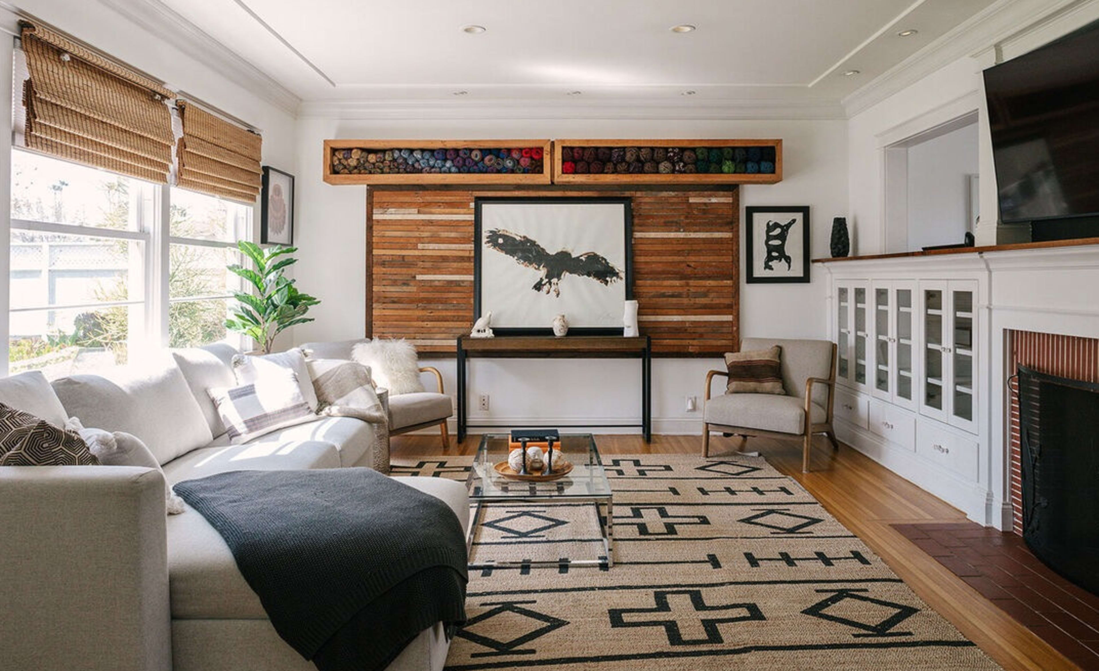 The living room features a light gray sectional sofa, a glass coffee table, and a wall adorned with wooden panels and artwork, alongside a potted plant and a television.
