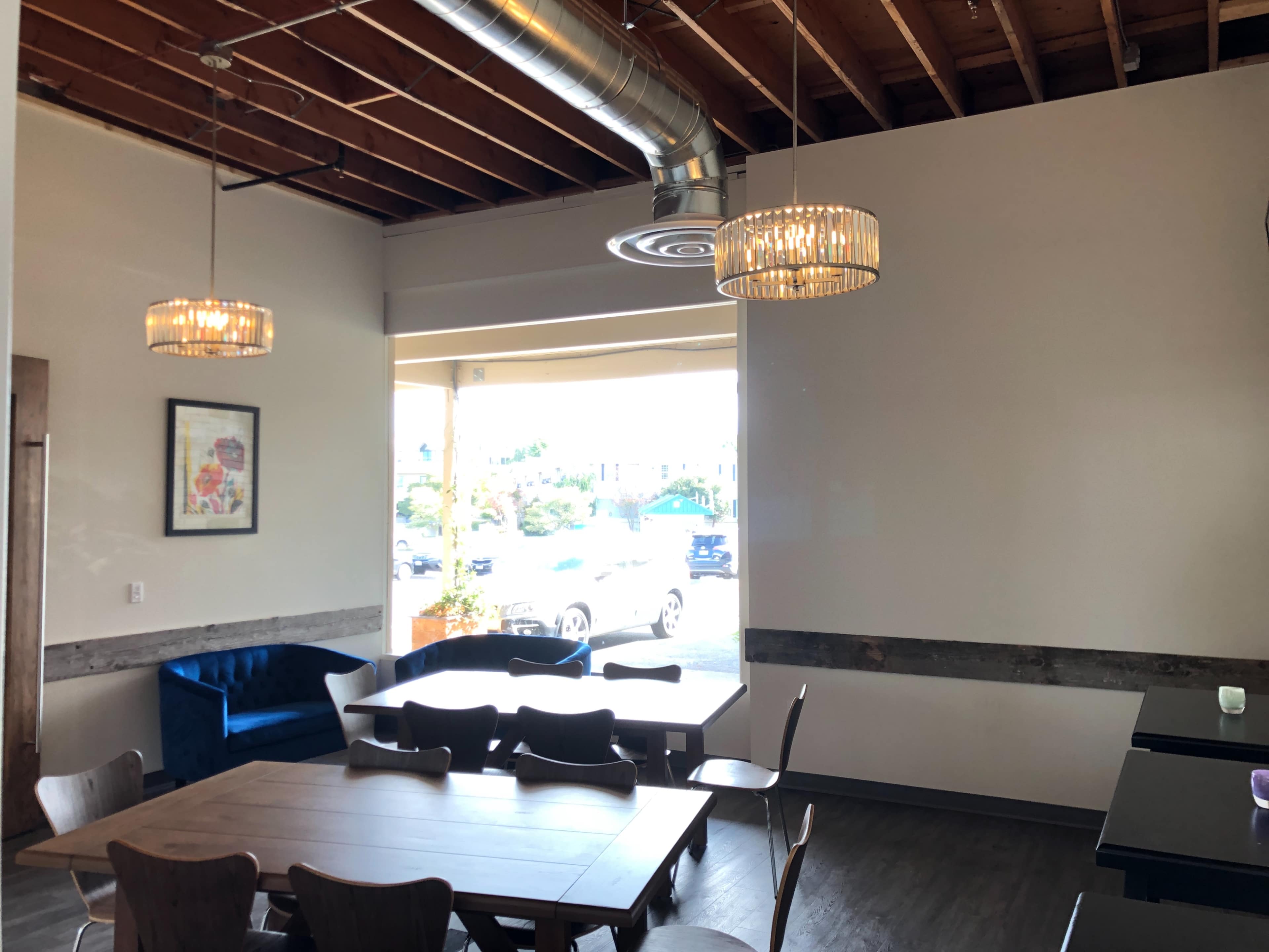The image shows a modern dining area with wooden tables, chairs, and two pendant lights, featuring large windows that provide natural light.