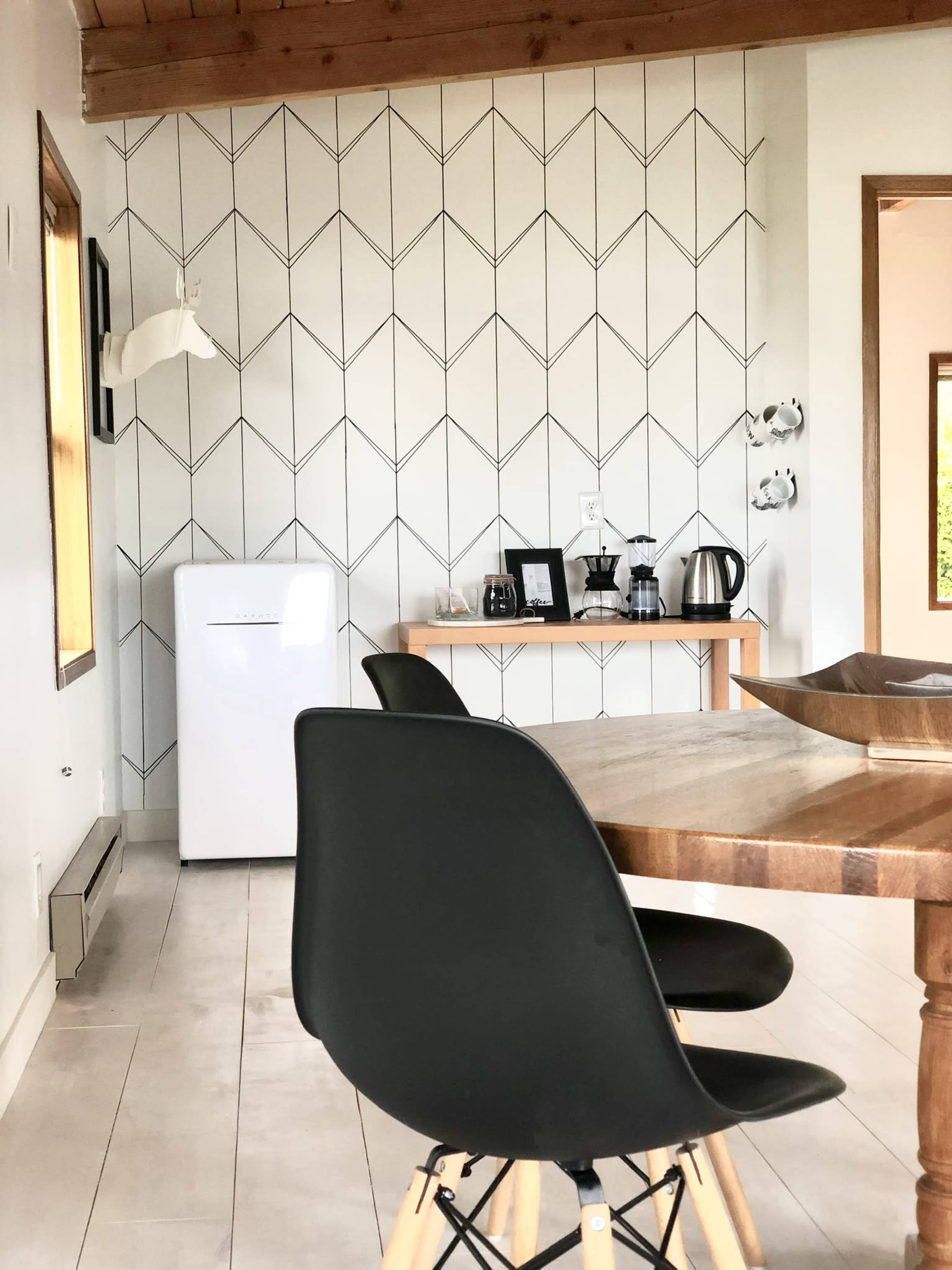 A modern kitchen area featuring a geometric-patterned wall, a white refrigerator, a wooden dining table with black chairs, and a countertop with various kitchen appliances.