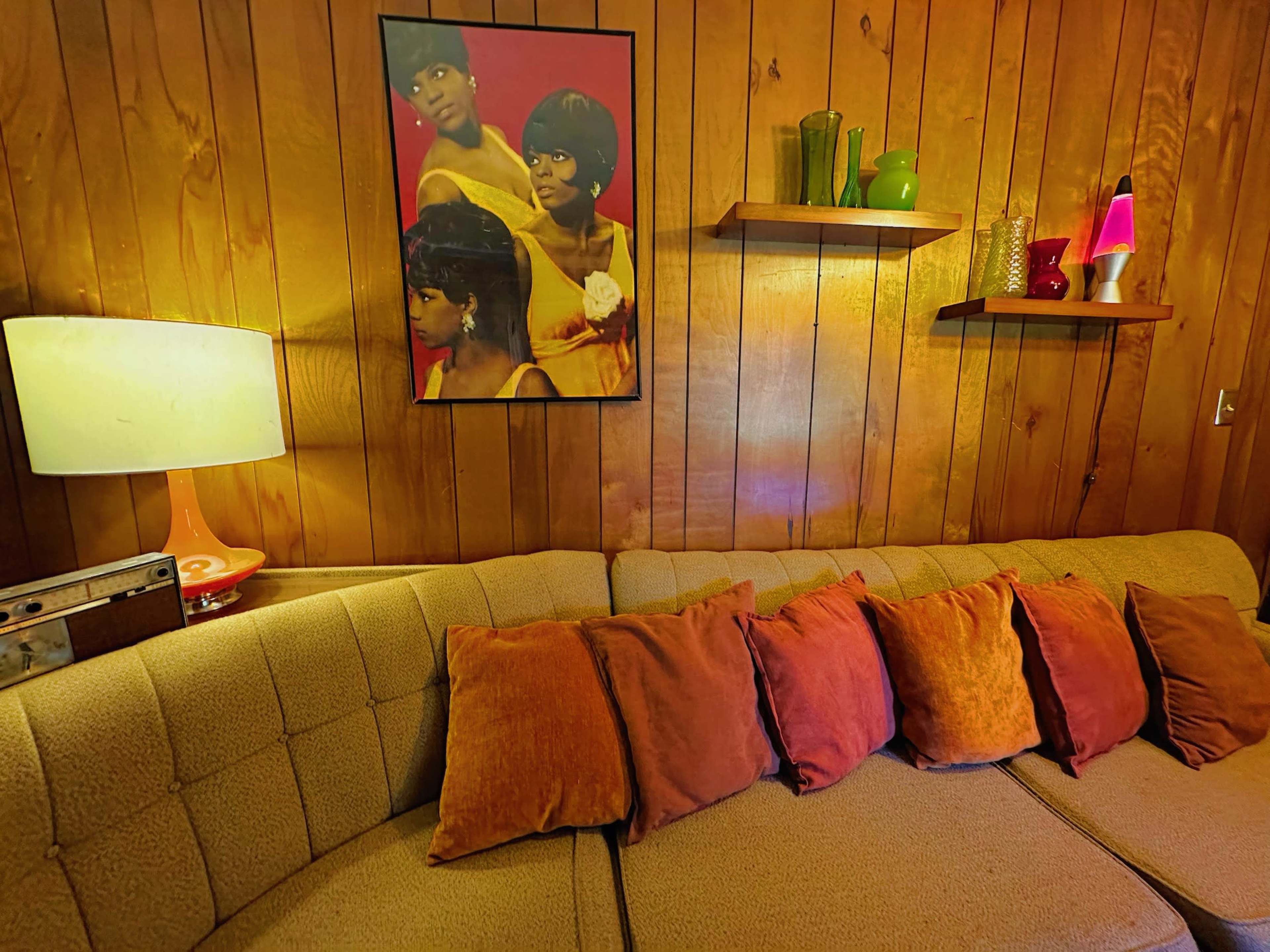 The image shows a living room with a retro-style couch adorned with orange and brown cushions, a framed artwork of three women on the wall, and decorative items on shelves along with a lamp.